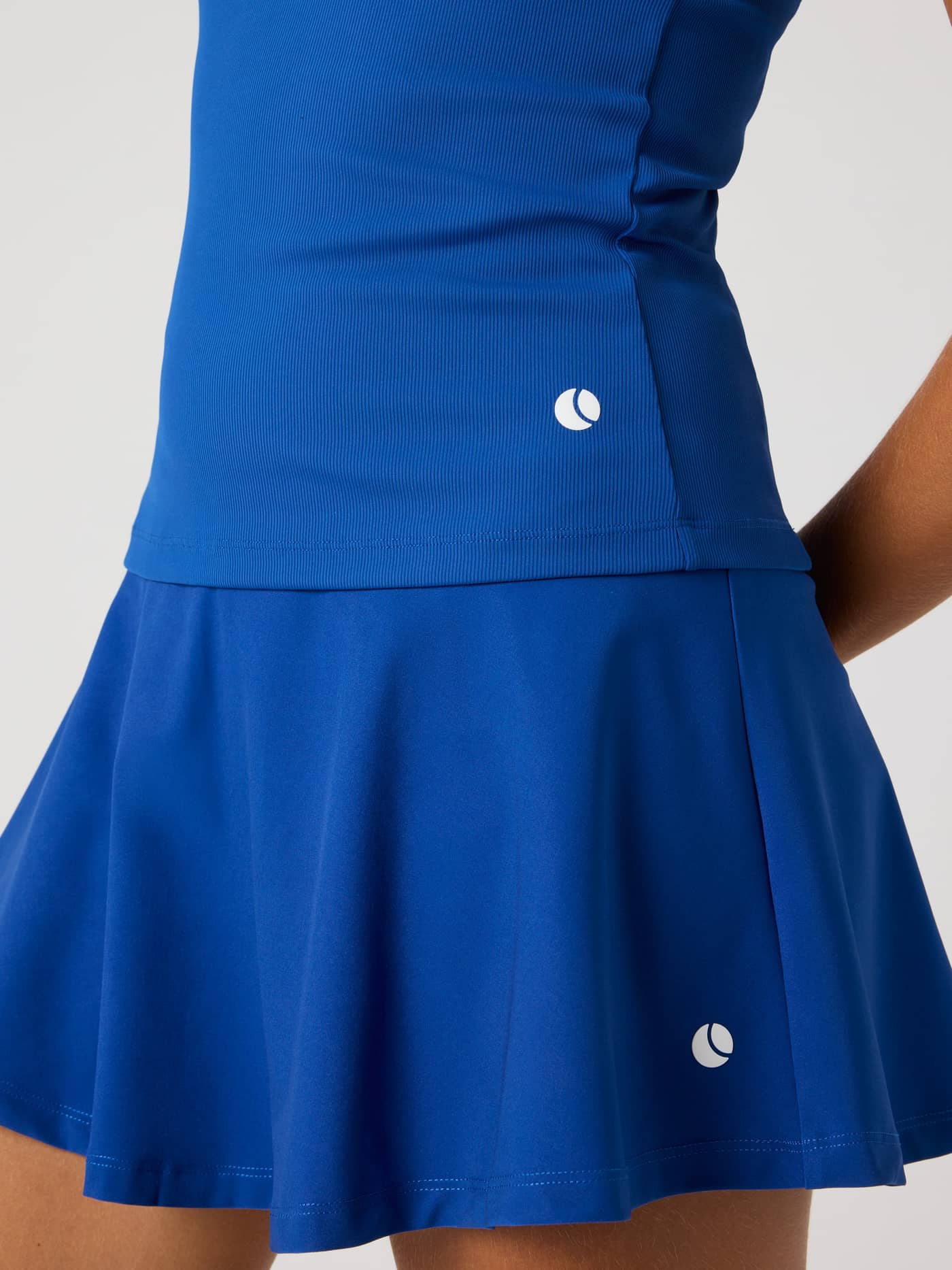 Närbild på ribbade Ace Pocket Ribbed Tank Top i Monaco Blue (Blå) med en vit tennisbollslogotyp på höften. - Image 5