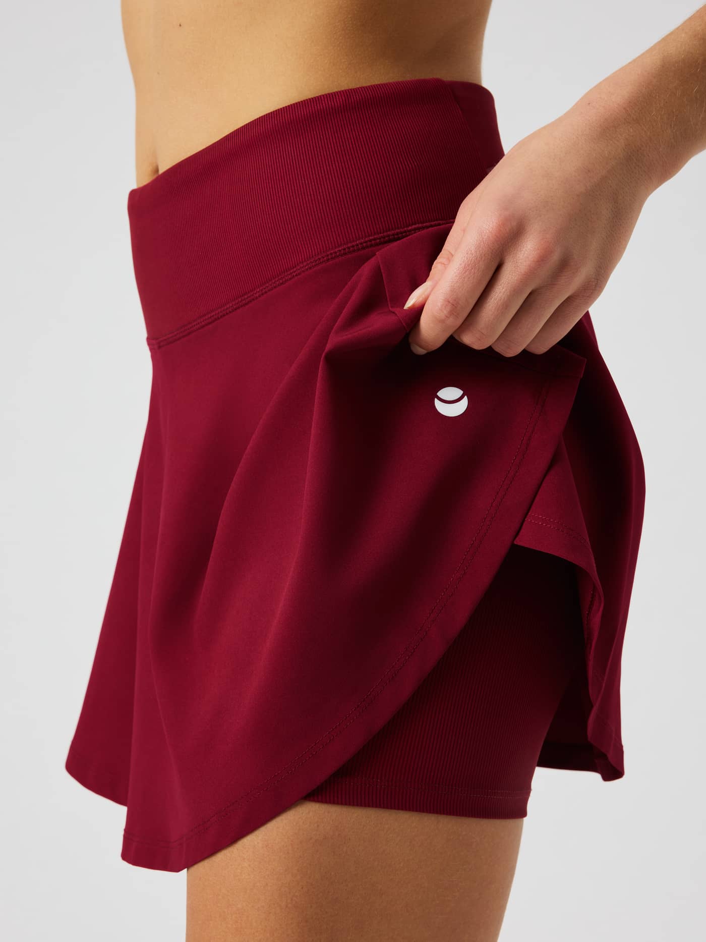 Close-up of the Ace Skirt Pocket in Cabernet (Red) highlighting the inner shorts and white tennis-ball logo. - Image 5
