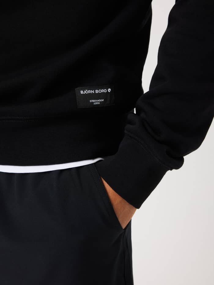 Close-up van de Centre Half Zip in Black Beauty (Zwart) met een geweven heuplabel en wit Björn Borg-logo. - Image 4