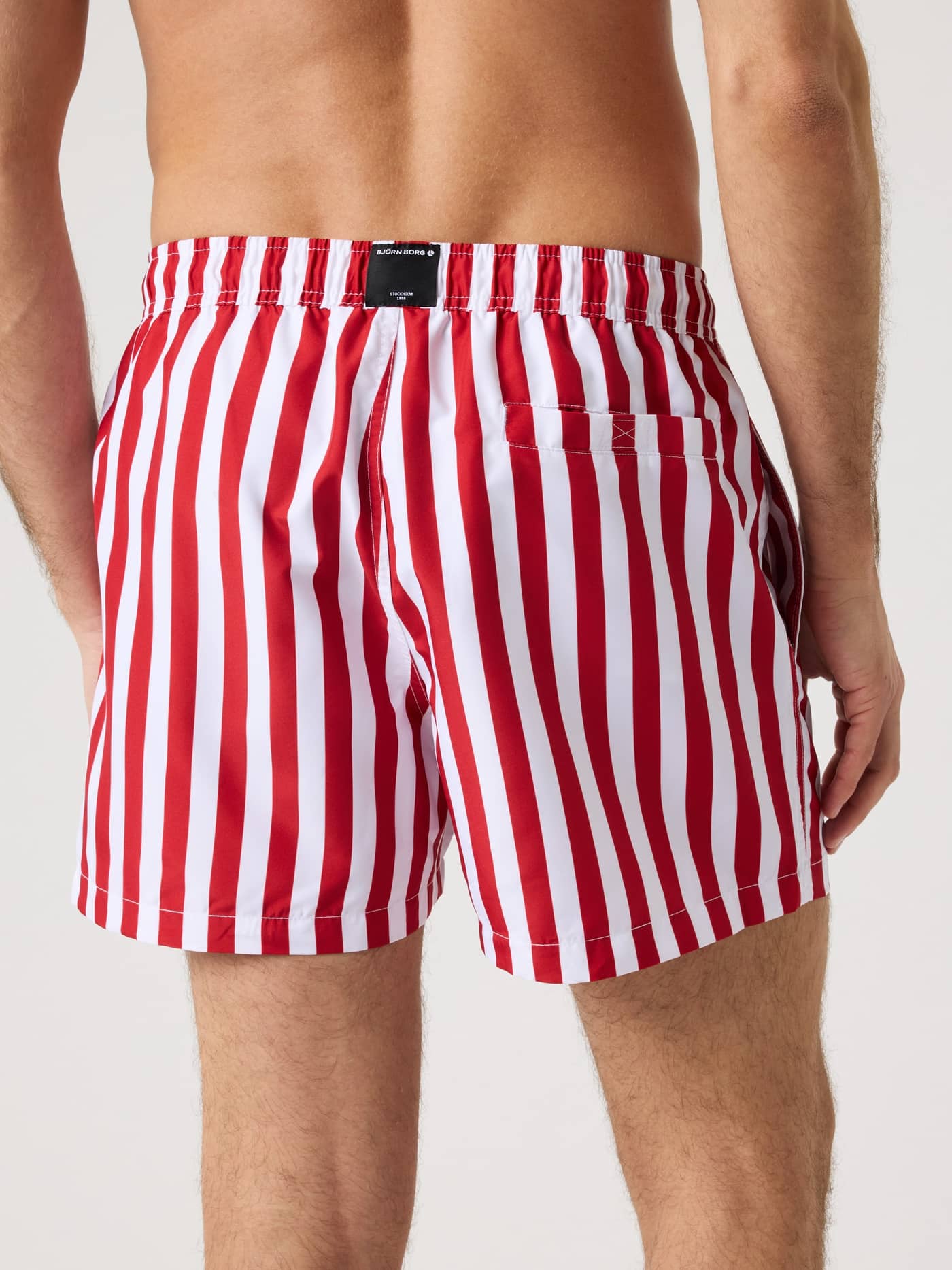 Back view of a man in striped Borg Print Swim Shorts in Parasol 3 (Red, White), made from quick-drying recycled polyester. - Image 2