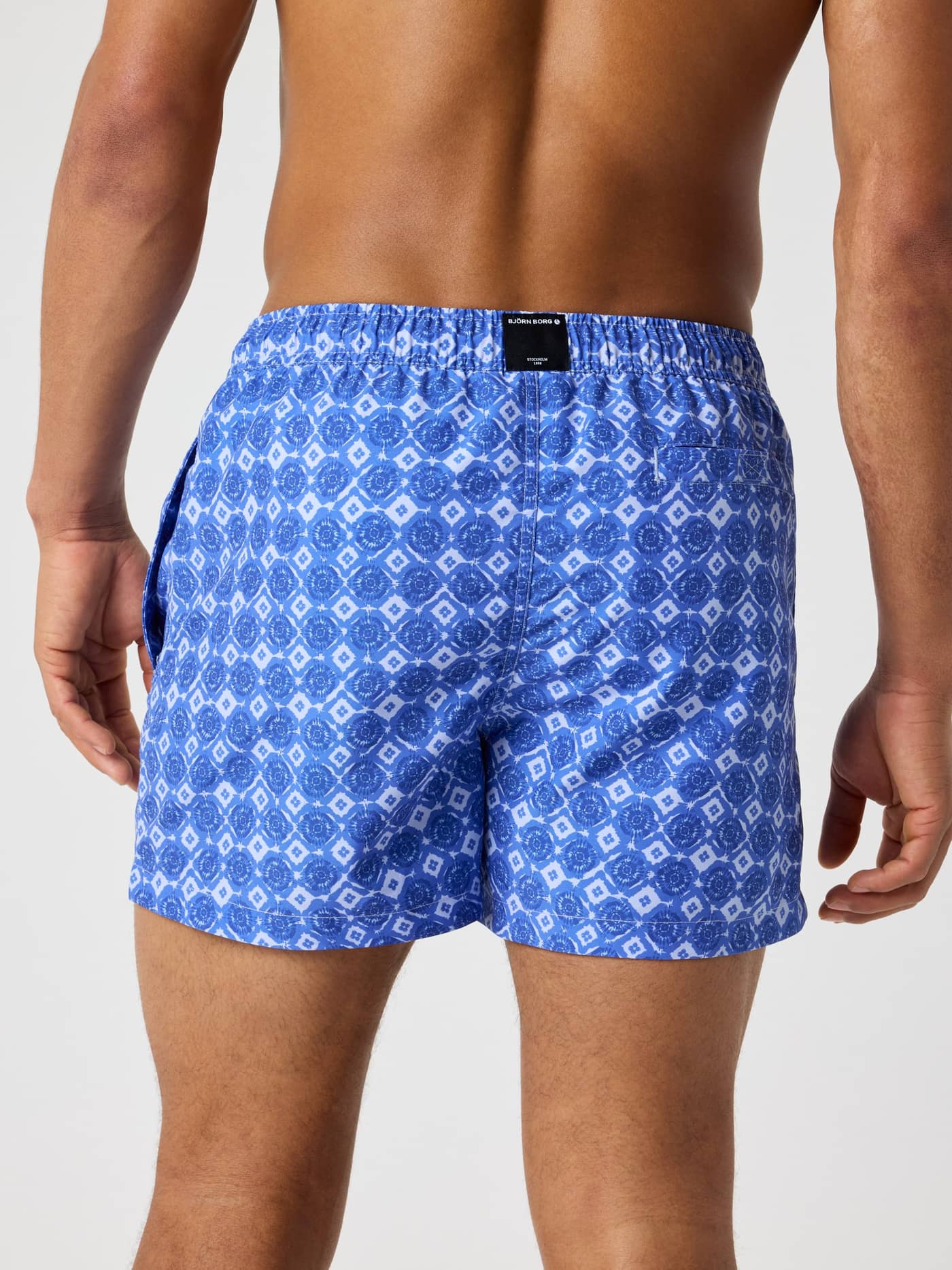 Back view of a man wearing Borg Print Swim Shorts in Sicily Swim 1 (Blue) with a geometric print and black logo tag. - Image 2