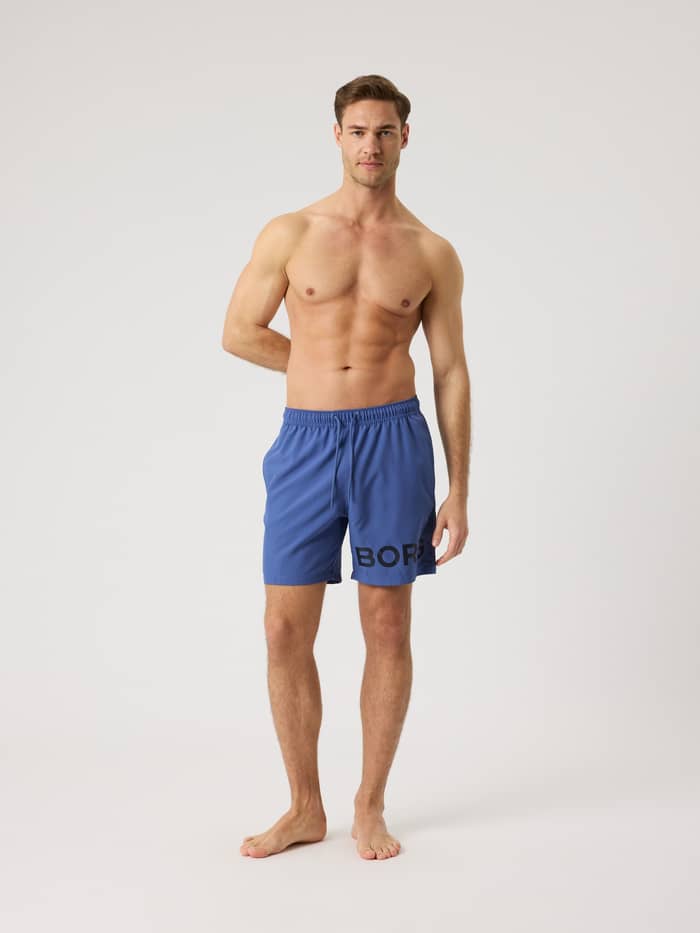 Volledig vooraanzicht van een man in de Borg Swim Shorts in Coastal Fjord (Blauw) met een opvallend logo en trekkoord. - Image 3