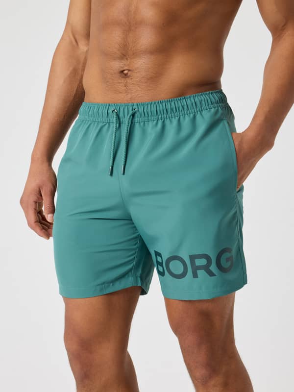 Front view of a man wearing the Borg Swim Shorts in Blue Spruce (Green), with a dark grey logo and functional drawcord.