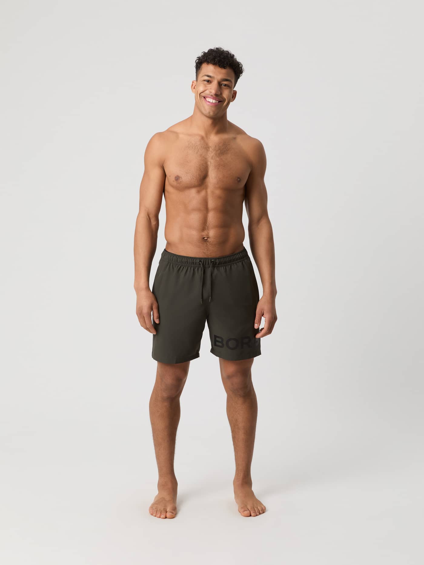 Mand i helfigur forfra iført funktionelle, hurtigtørrende Borg Swim Shorts i Peat (Grøn) med et stort, sort logo. - Image 3