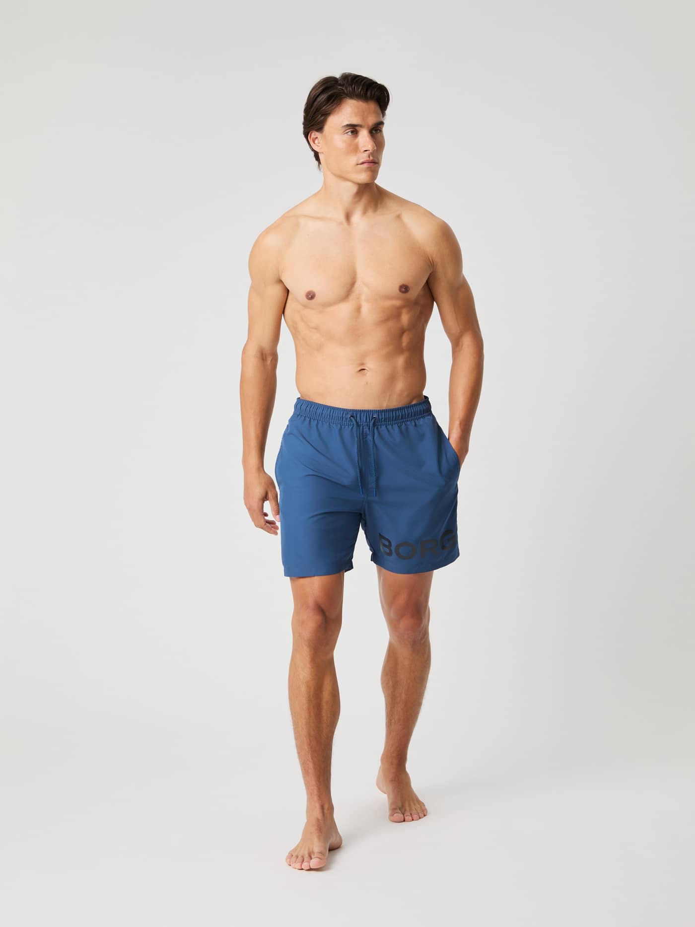 Full-body front view of a man wearing functional Borg Swim Shorts in Sargasso Sea (Blue) with a black logo on the leg. - Image 1