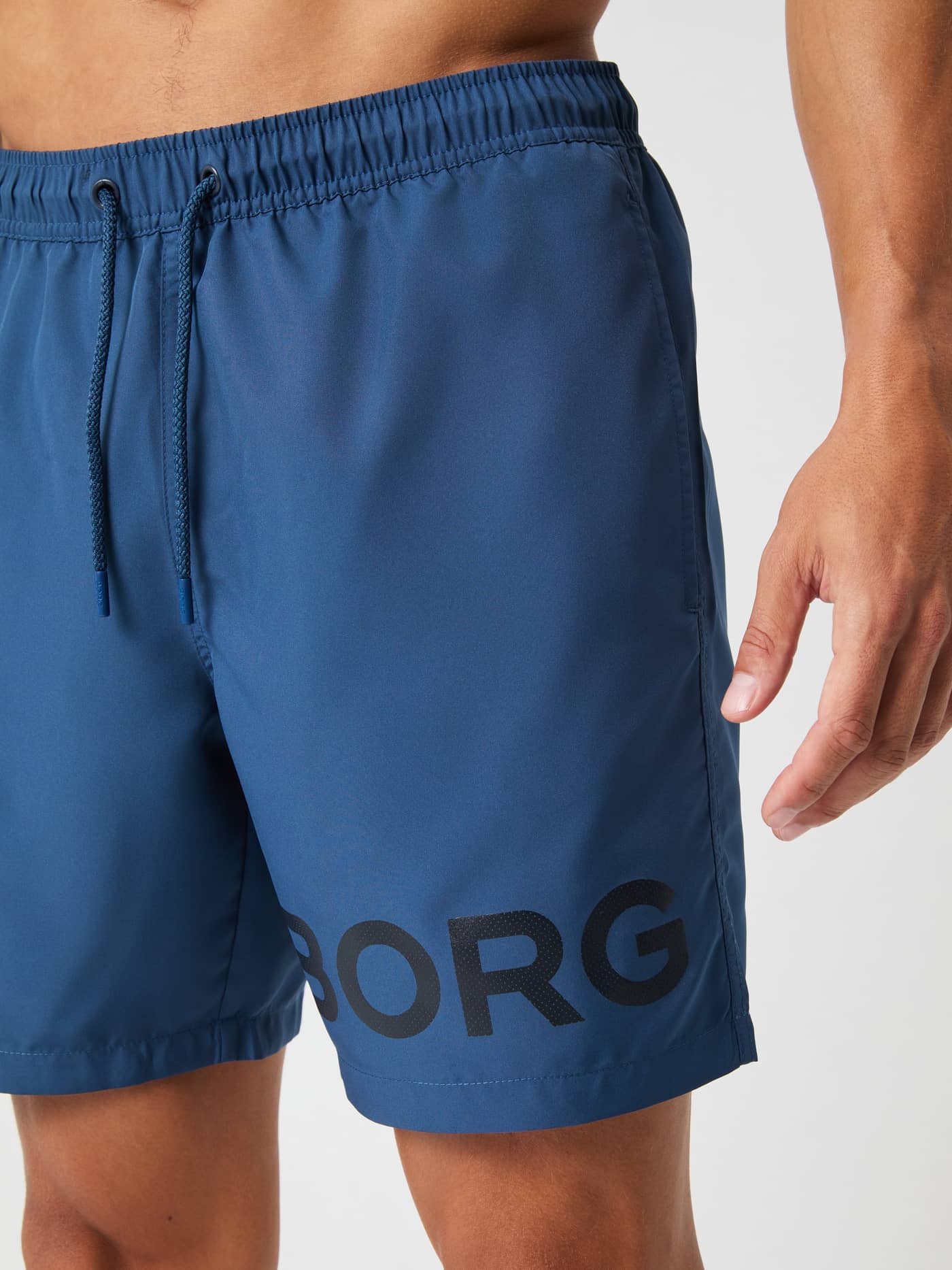Close-up of a man wearing Borg Swim Shorts in Sargasso Sea (Blue) with a bold black logo and adjustable drawstrings. - Image 4