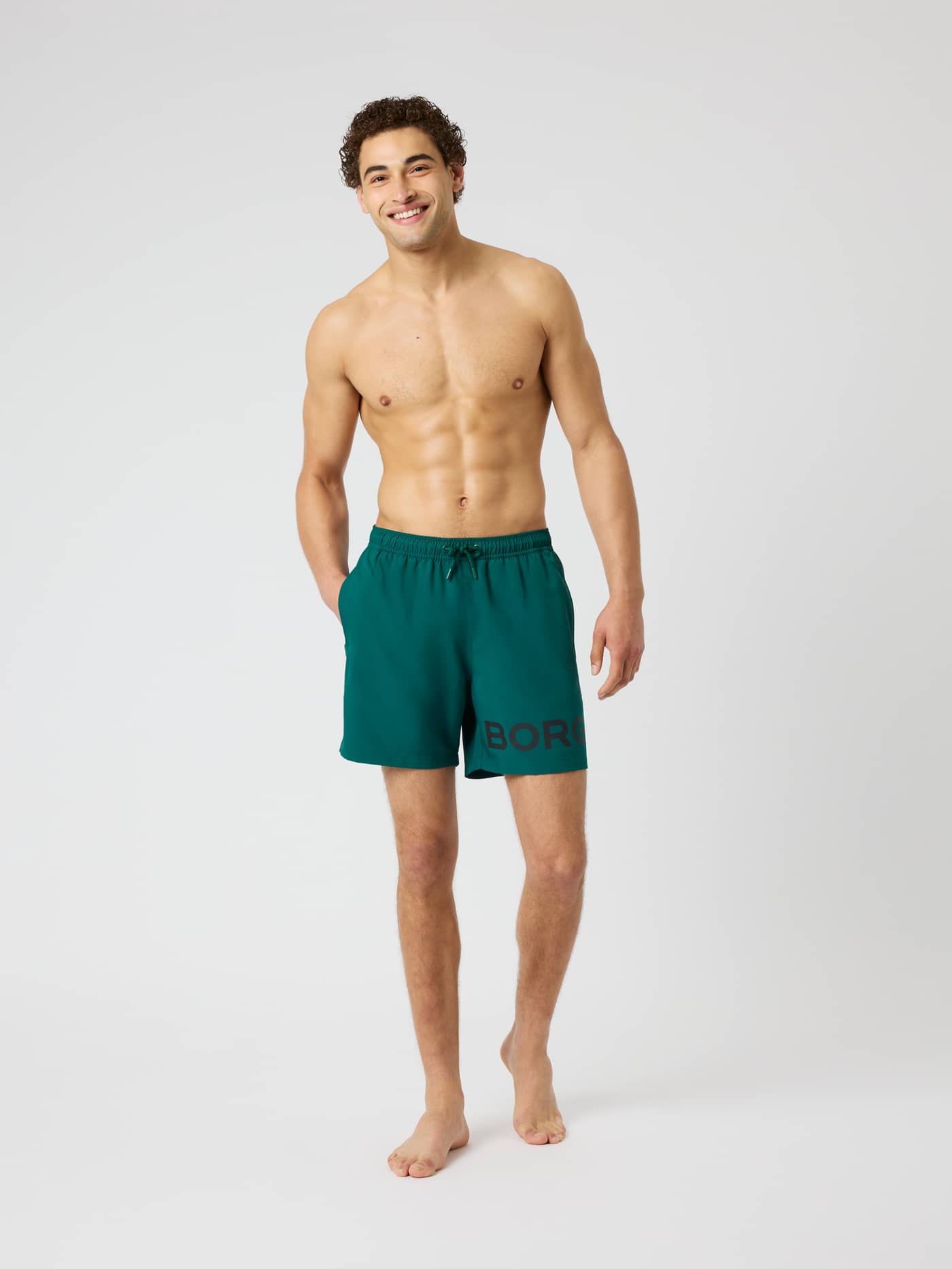Front view of a man wearing Borg Swim Shorts in Rain Forest (Green) featuring a drawstring waist and black logo on the leg. - Image 3