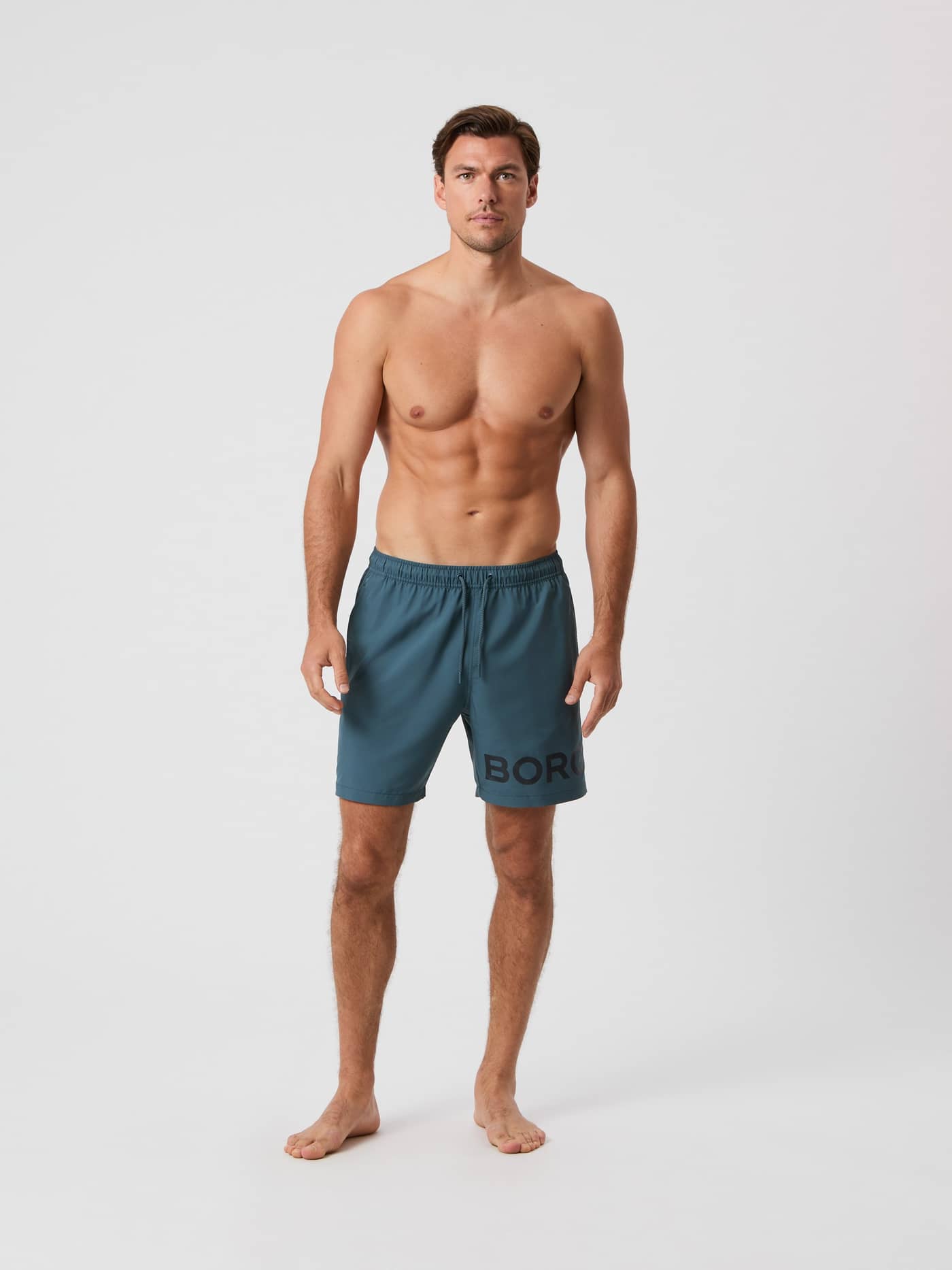 Full-body front view of a man wearing the Orion Blue (Blue) Borg Swim Shorts featuring a bold logo on the leg. - Image 1
