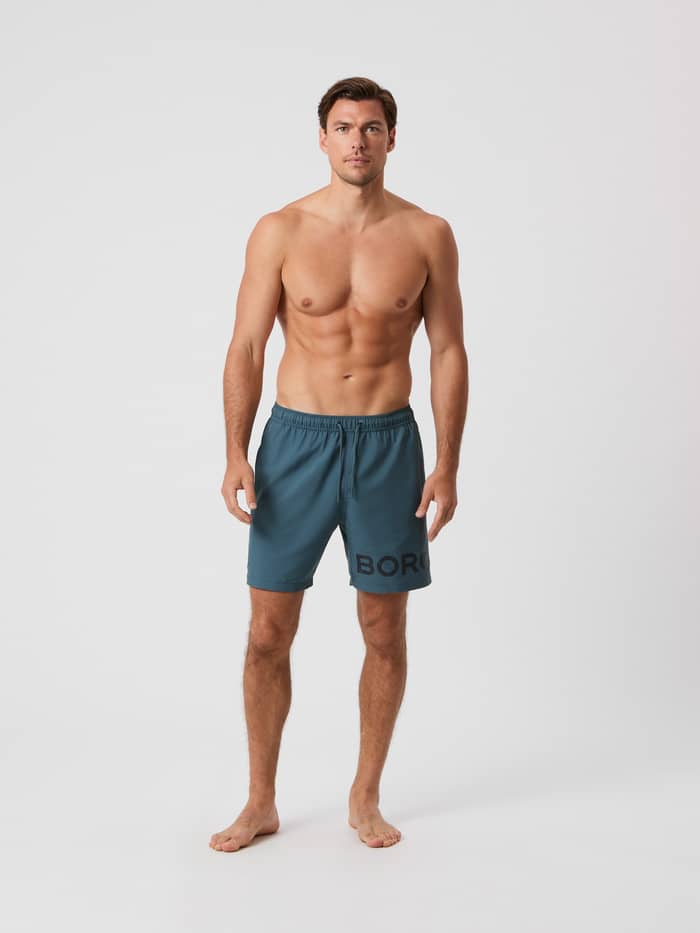 Helkropp framifrån av en man i Orion Blue (Blå) Borg Swim Shorts med en stor mörk logotyp på benet.