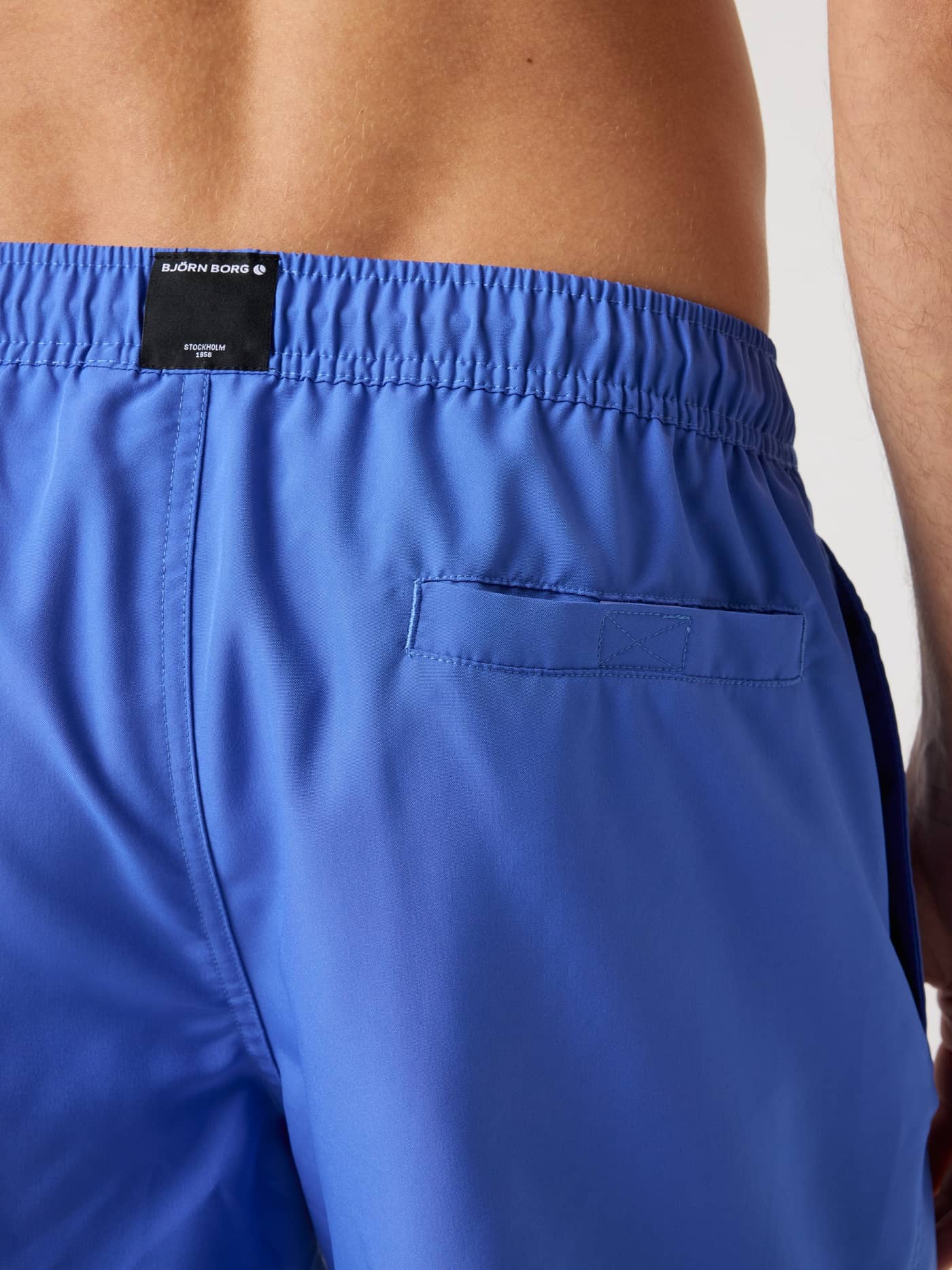 Detailed back view of a man wearing Björn Borg Solid Swim Shorts in Amparo Blue (Blue) with a black logo and Velcro pocket. - Image 4