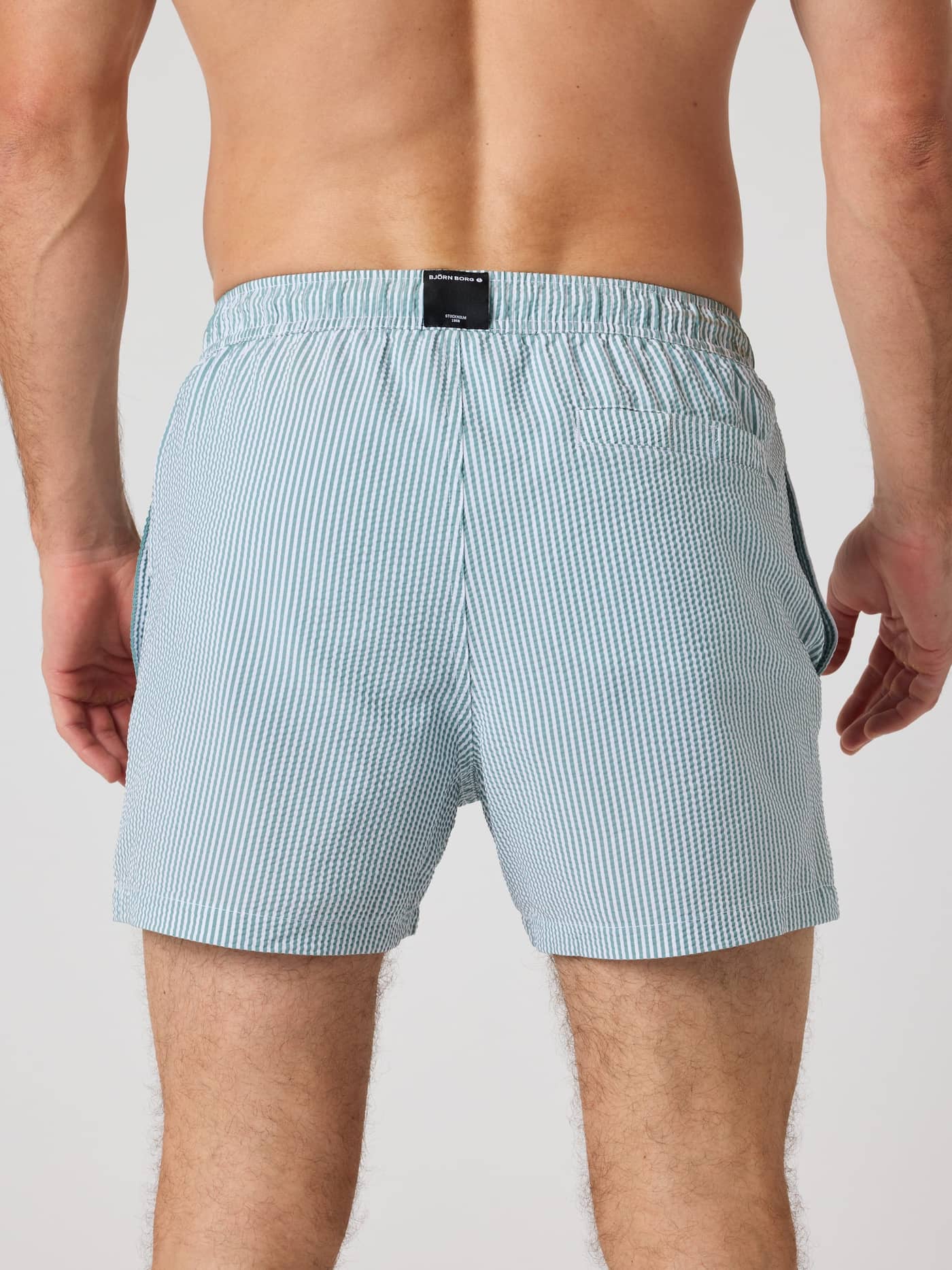 Back view of a man wearing the striped Borg Seersucker Swim Shorts in Searstripe 5 (Blue) with a black logo label. - Image 2