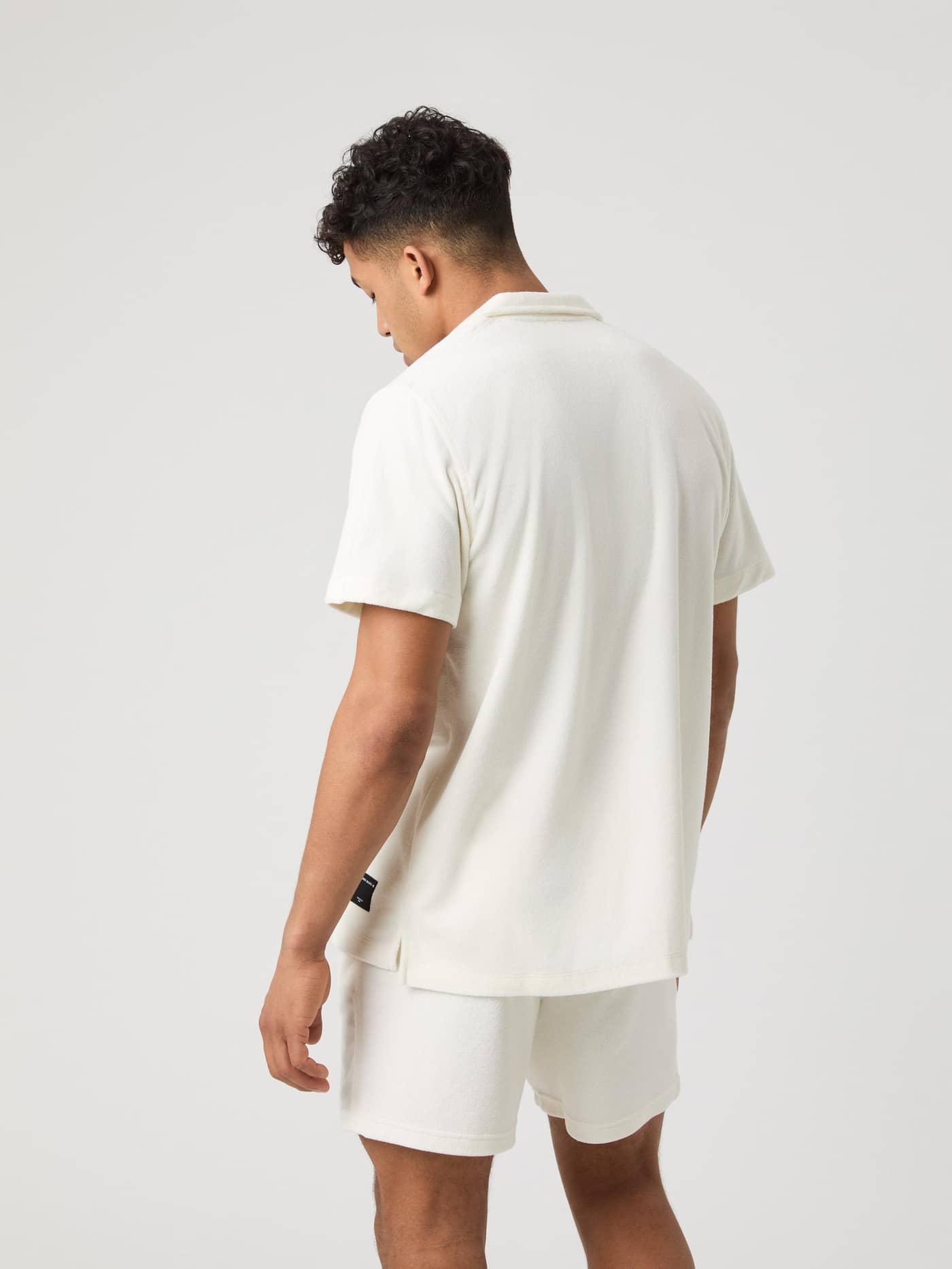 Rückansicht eines Mannes im Borg Toweling Pool Shirt in Egret (Weiß) mit schwarzem Logo-Label und Seitenschlitzen. - Image 2