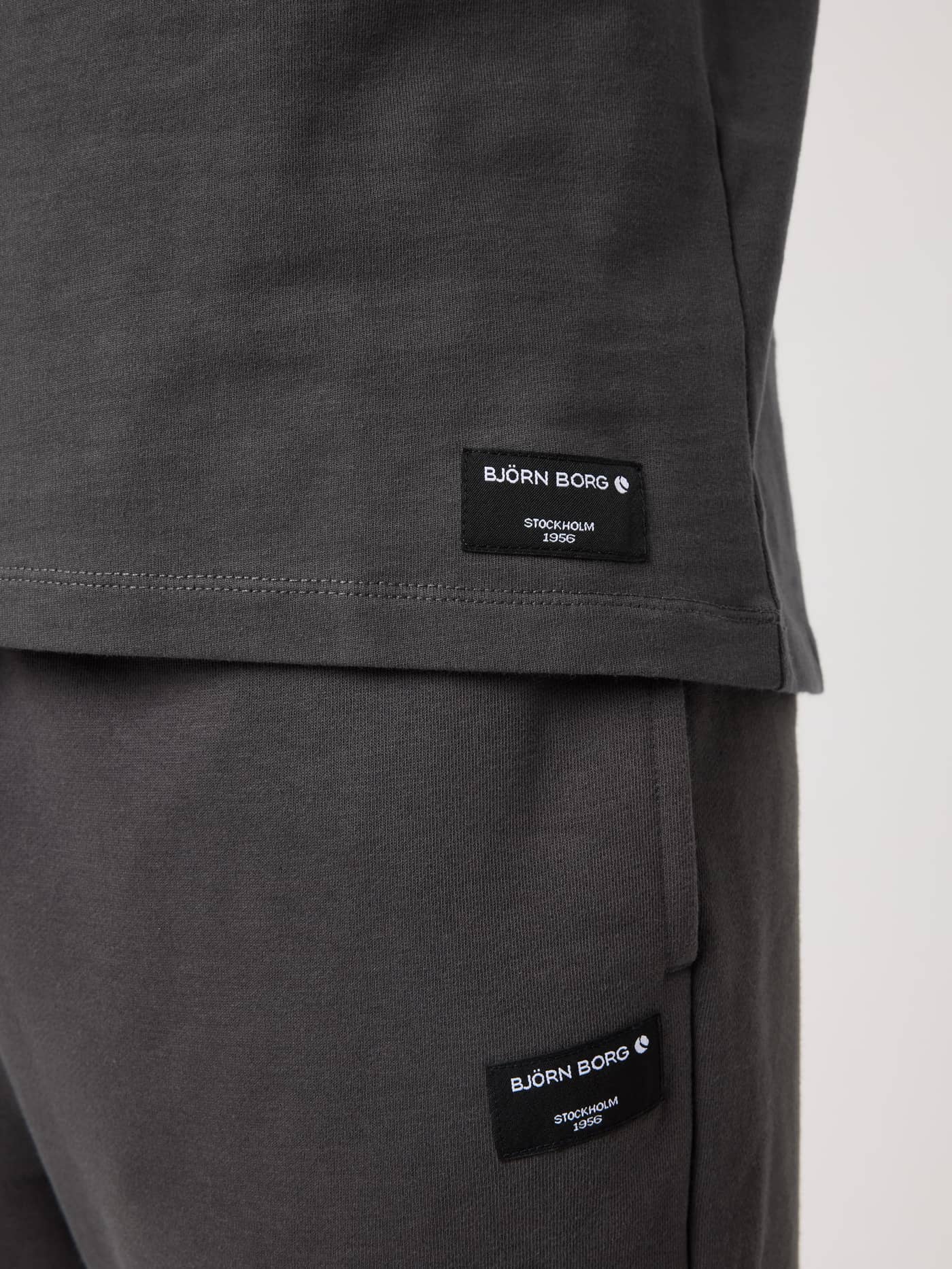 Close-up of the Centre High Neck T-Shirt in Asphalt (Grey) featuring a black logo label on the side hem. - Image 4