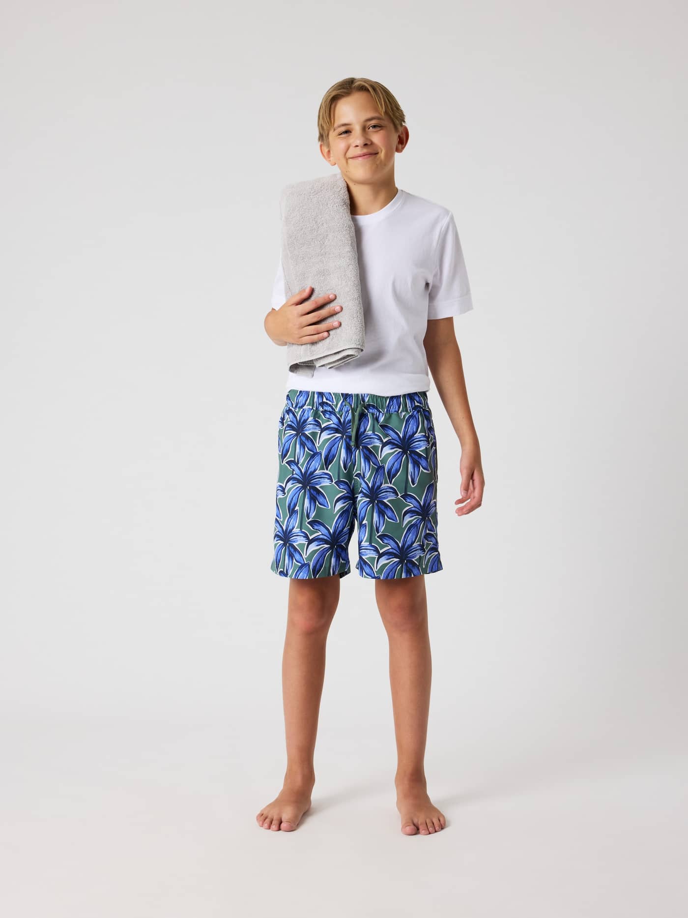 Borg Print Swim Shorts för barn i Palms On Palms 1 (Blå/Grön) med palmtryck sedda i helfigur framifrån på en pojke. - Image 1
