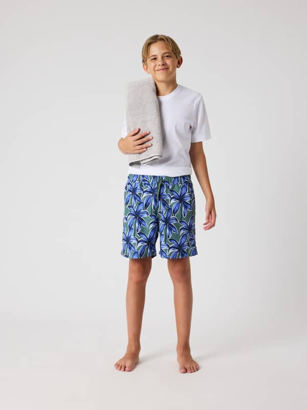 Borg Print Swim Shorts til kids i Palms On Palms 1 (Blå/Grøn) med palmetræsprint på en dreng i fuld figur set forfra.