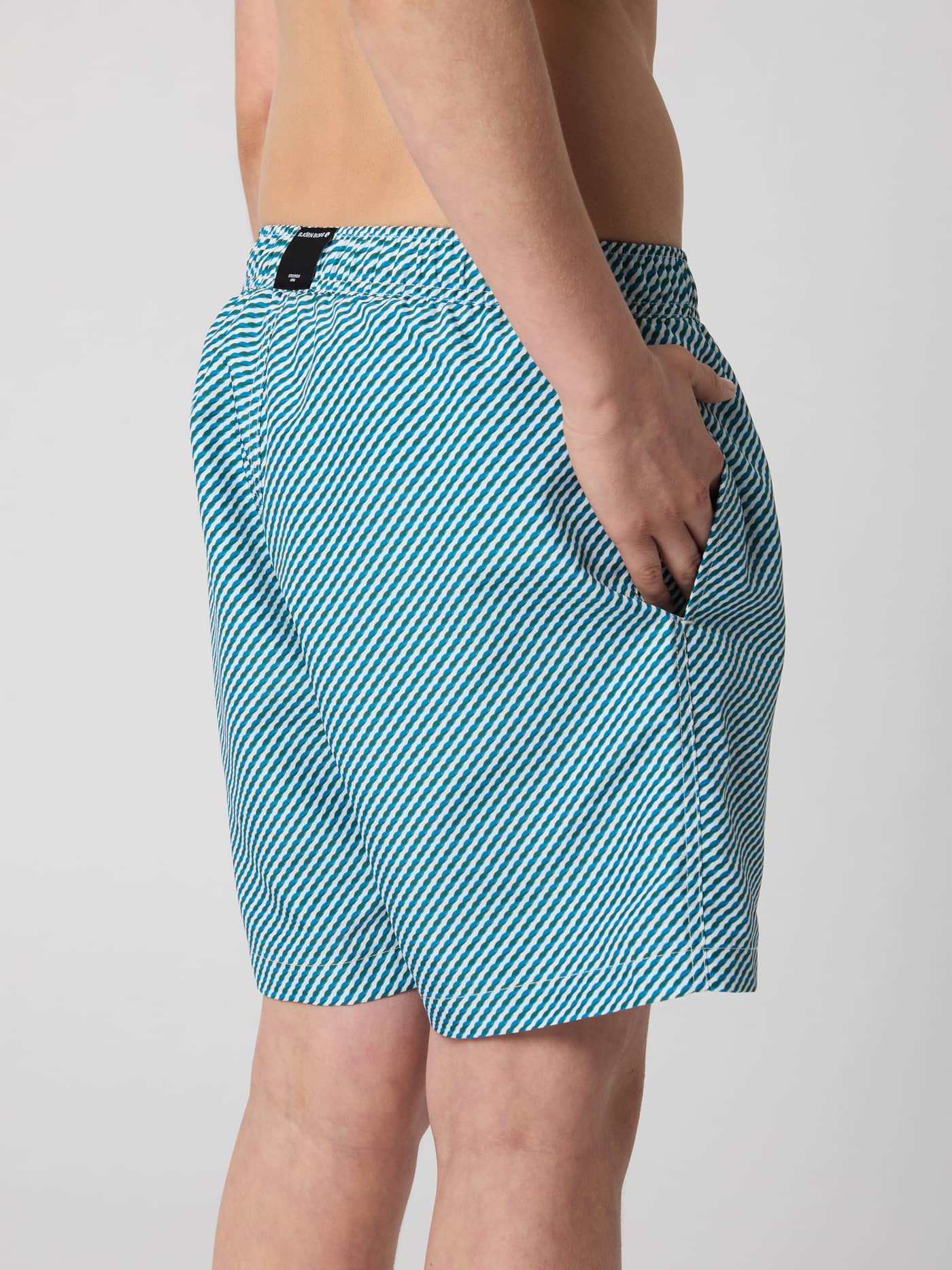 Side view of a kid wearing Borg Print Swim Shorts in Candy Stripe 2 (Blue) with a striped pattern and a black logo tag. - Image 3