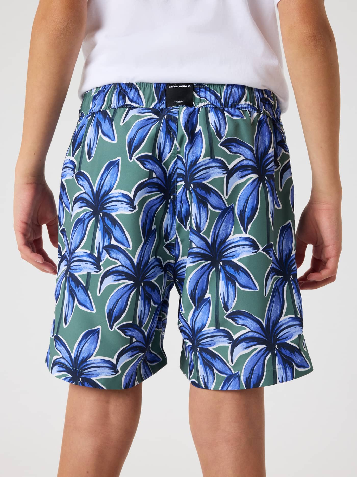 Back view of a model wearing kids Borg Print Swim Shorts in Palms On Palms 1 (Blue Green) with an all-over palm print. - Image 3