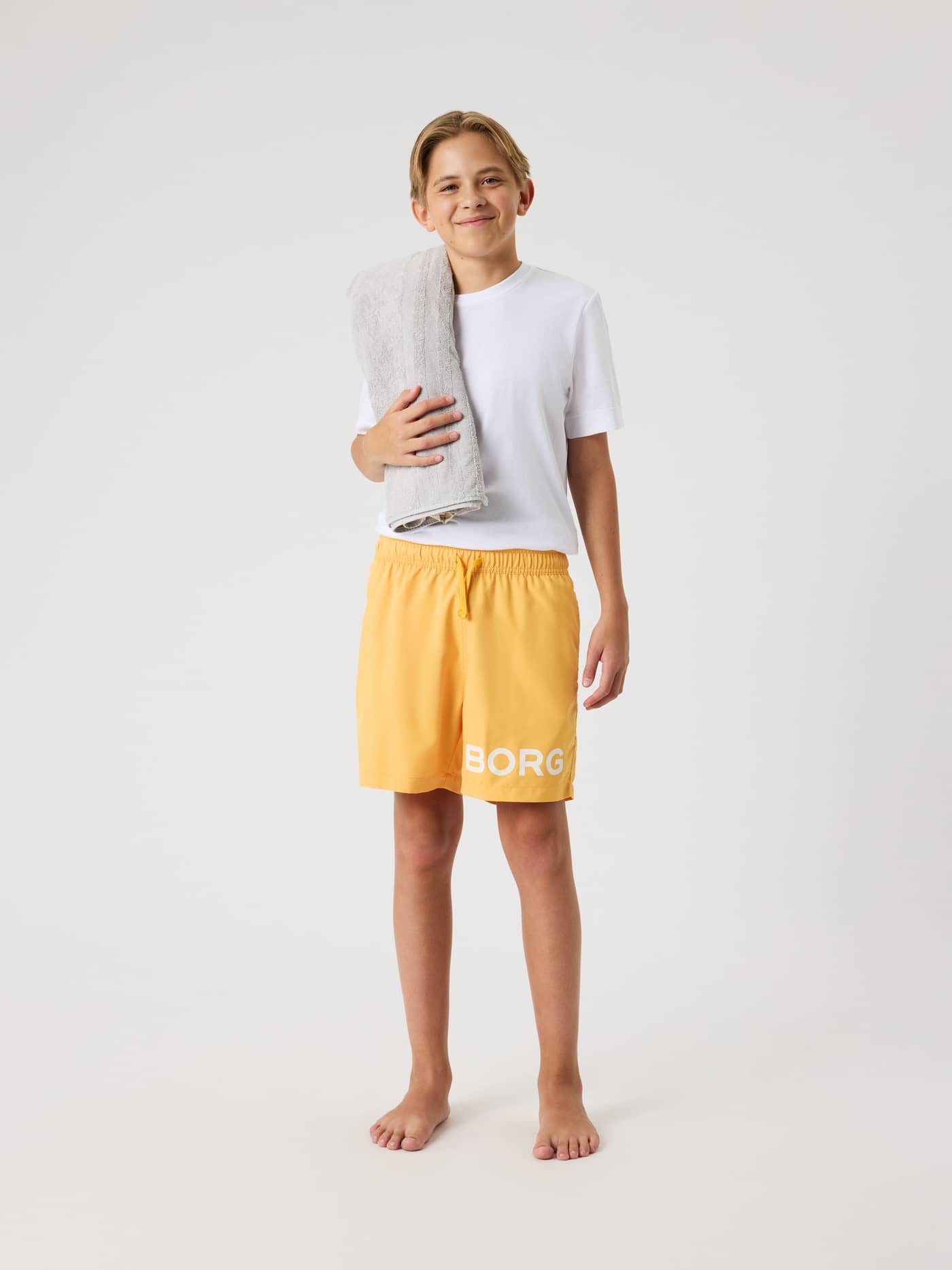 Borg Swim Shorts til børn i Marigold (Gul) med hvidt logo og almindelig pasform, vist forfra med et gråt håndklæde. - Image 3