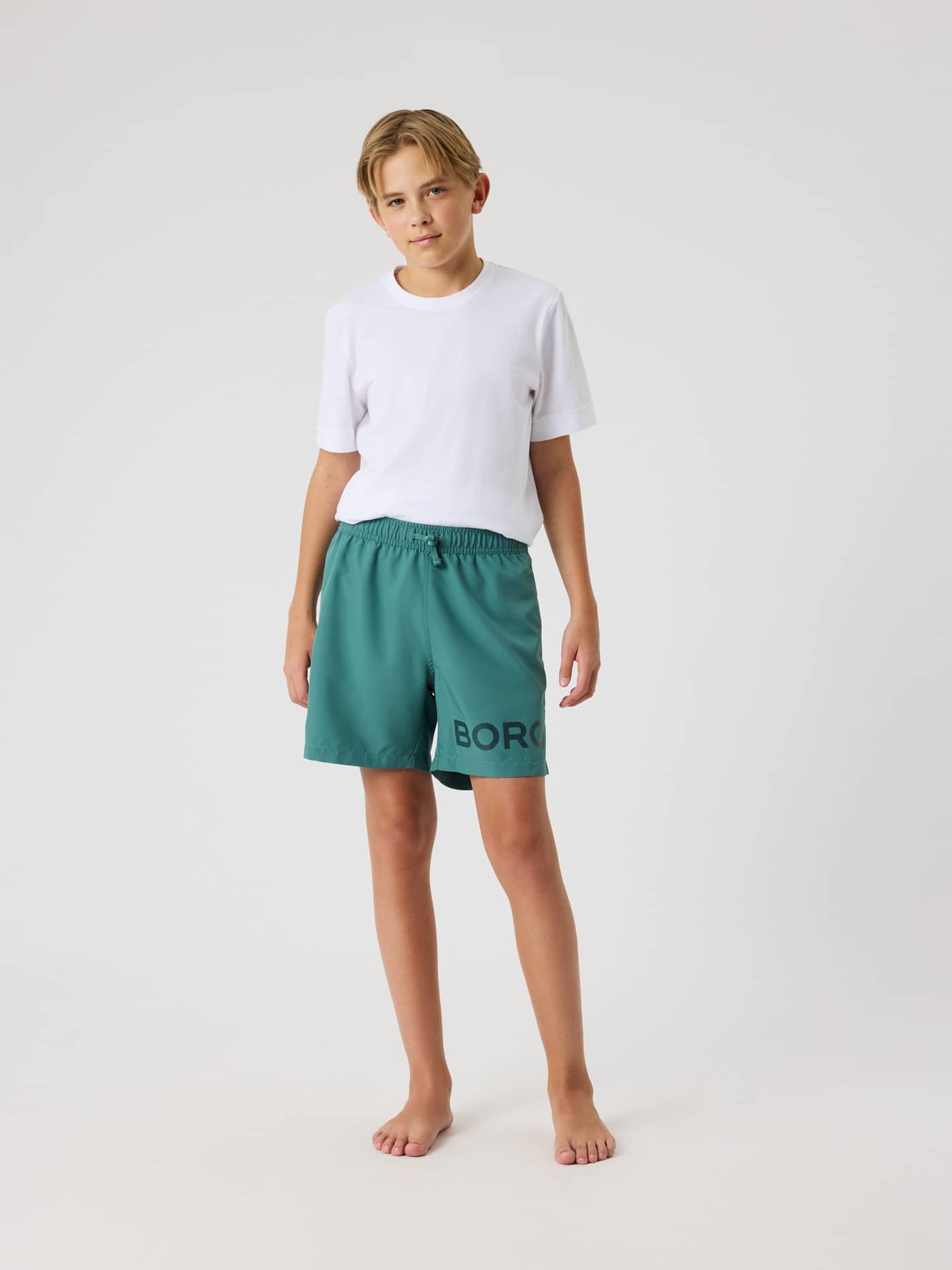 Full-body front view of a model wearing the kids' Borg Swim Shorts in Blue Spruce (Green) with a regular fit and bold logo. - Image 3