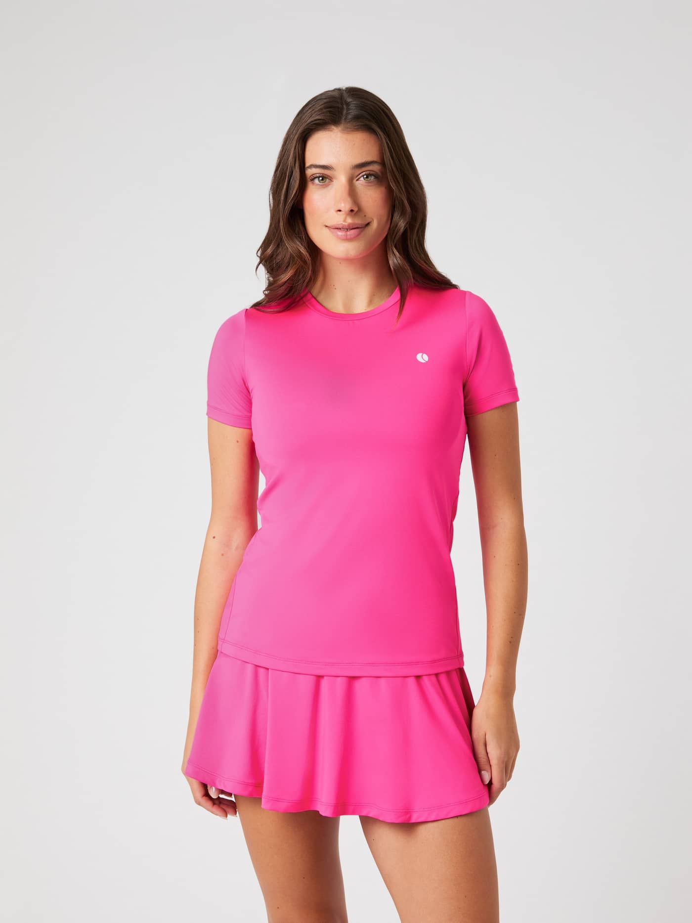 Front view of a woman wearing the bright pink Björn Borg Ace Slim T-shirt and matching skirt against a grey background. - Image 1