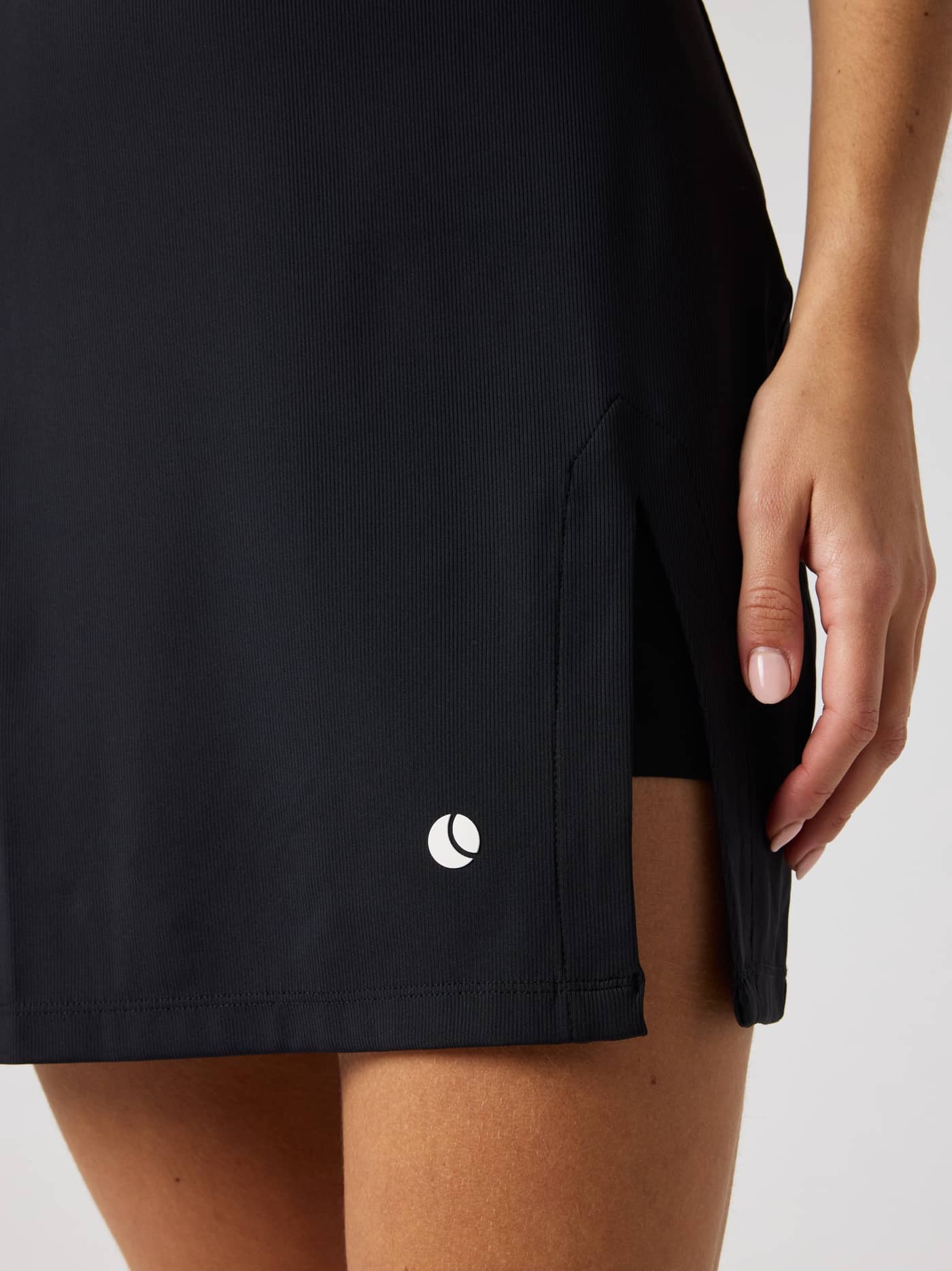 Ace Ribbed Dress in Black Beauty (Zwart) in een detailopname met zijsplit en wit tennisballenlogo voor tennis. - Image 4