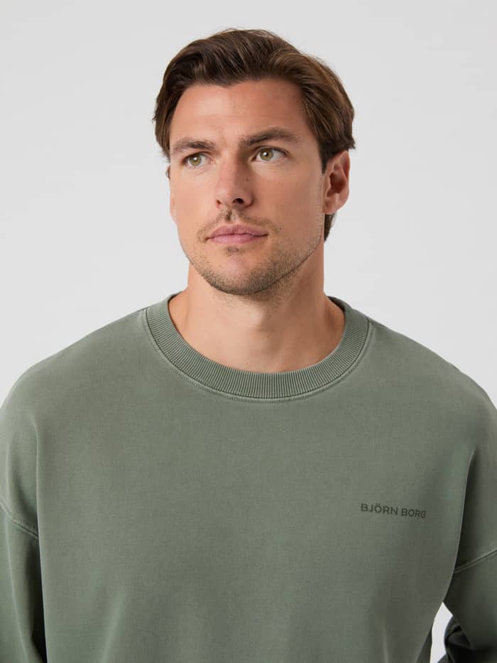 Borg Oversized Washed Sweatshirt in Thyme (Groen) met een donker geborduurd logo, gedragen door een man in close-up. - Image 5