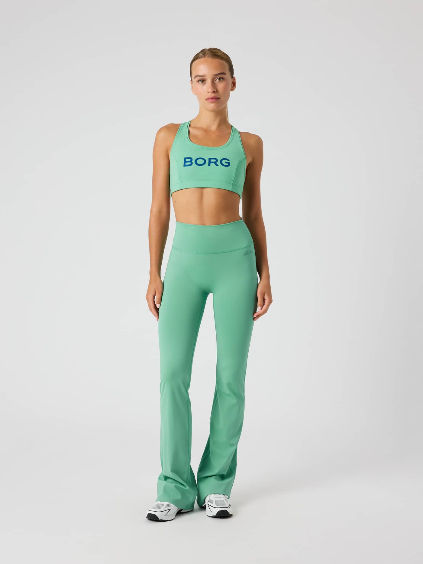 Full-body front view of a woman wearing the green Borg Mid Support Sports Bra with a blue logo for a stylish training fit. - Image 3