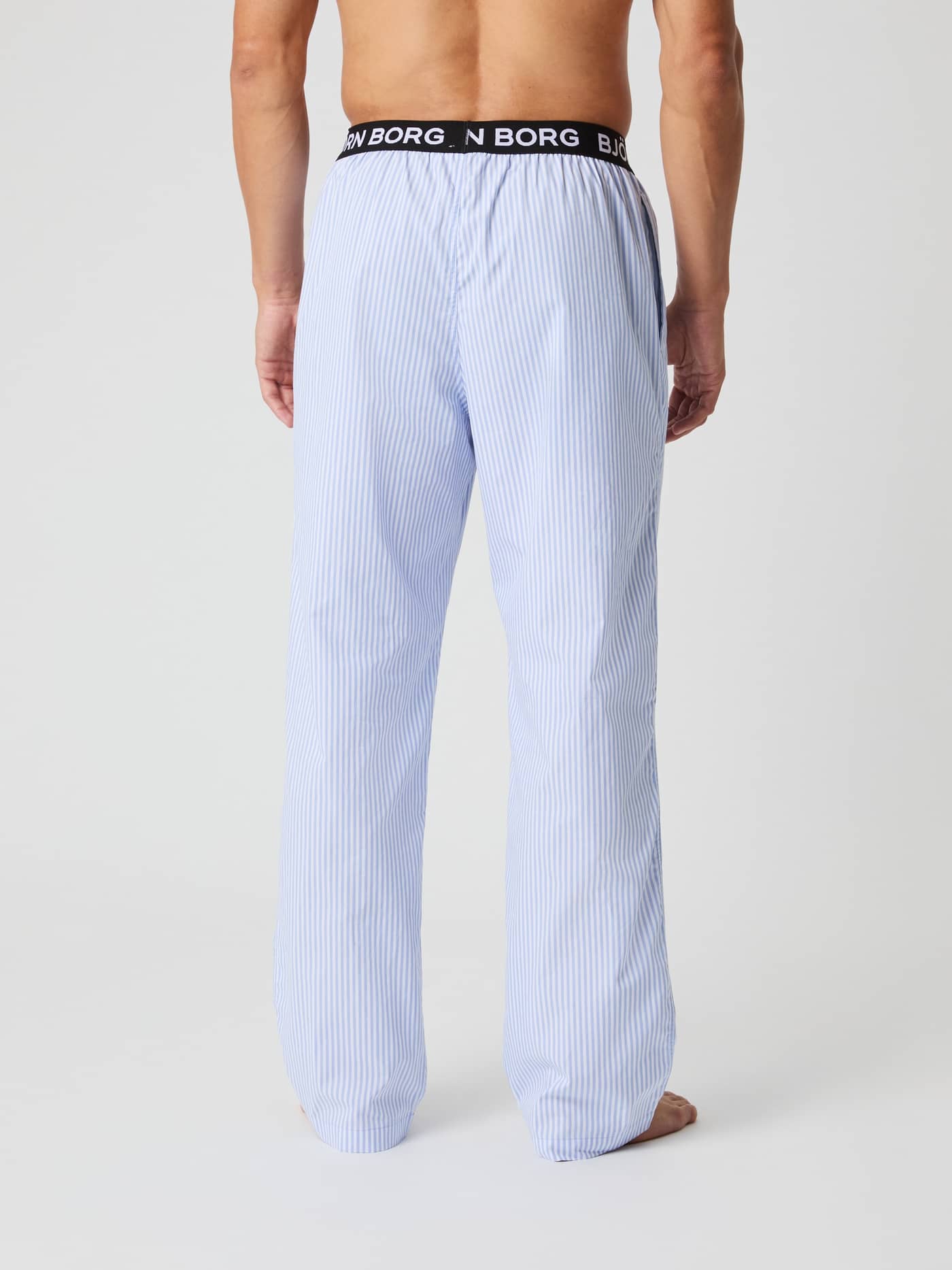 Back view of a man wearing striped Core Woven Logo Pyjama Pants in Fresh Stripe 1 (Blue) with a black logo waistband. - Image 3