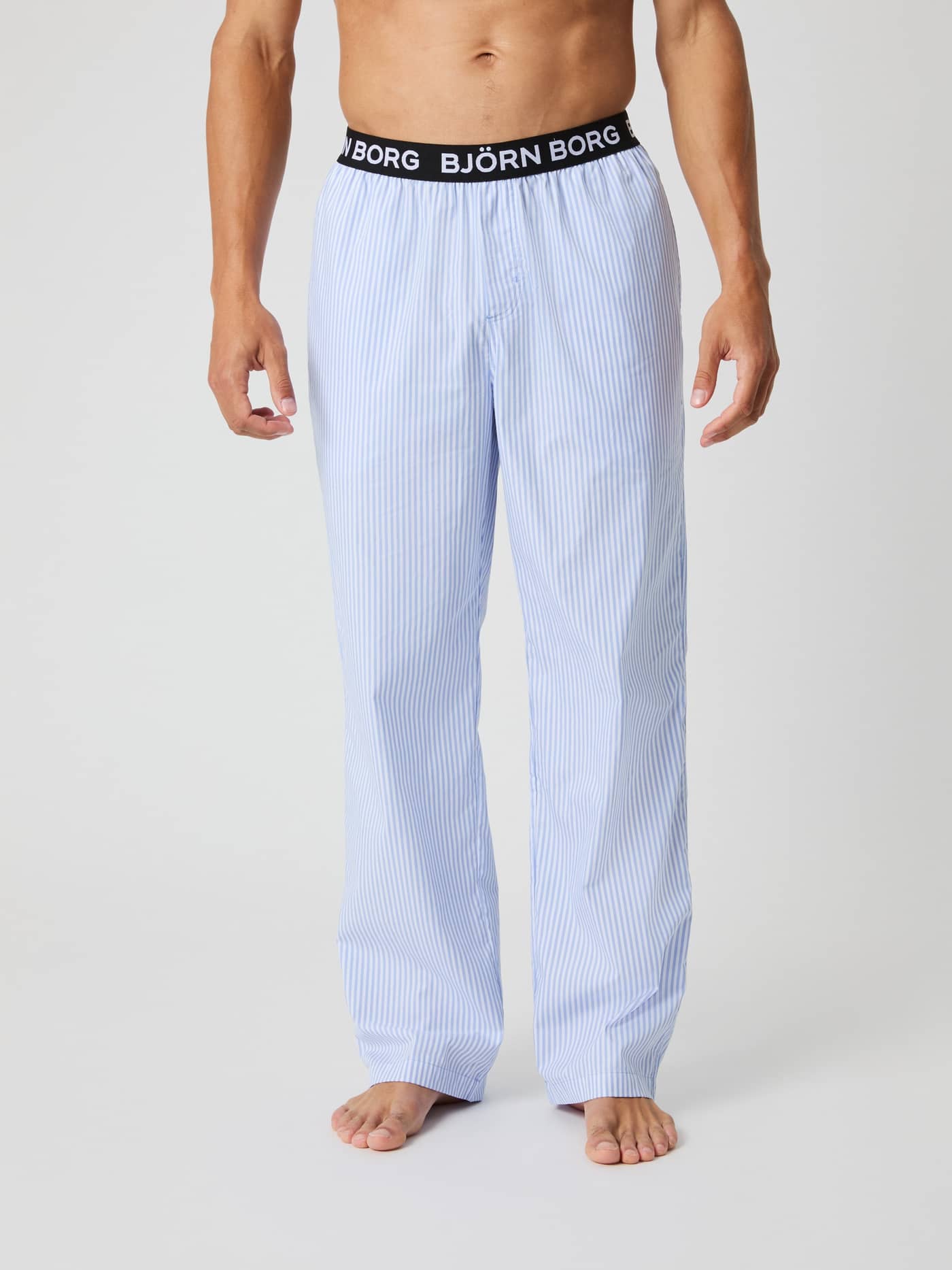 Front view of a man in Core Woven Logo Pyjama Pants in Fresh Stripe 1 (Blue) with vertical stripes and a logo waistband. - Image 2