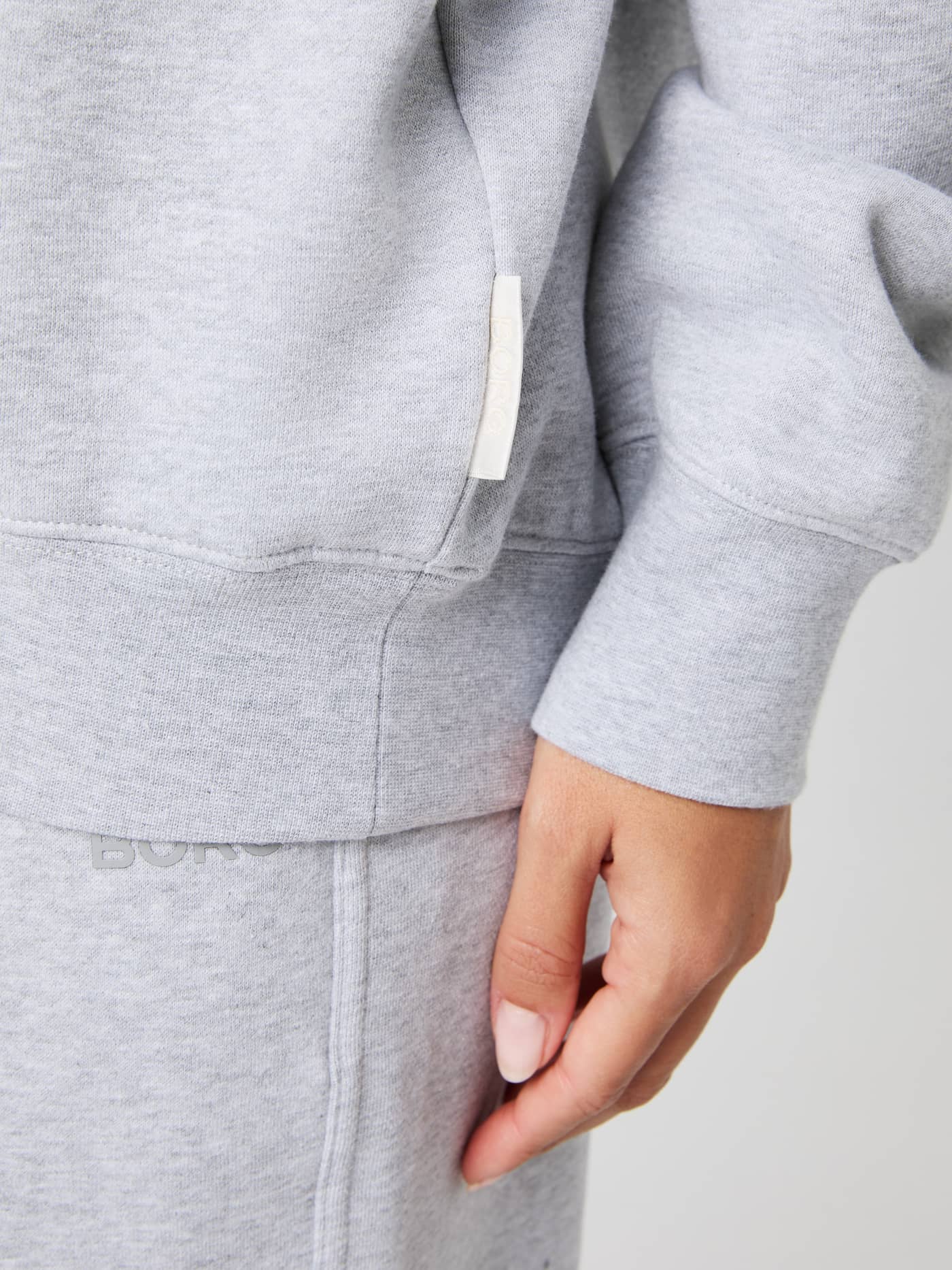 Close-up of the women's light grey melange Studio Oversized Hoodie, detailing the ribbed cuff, hem, and side seam label. - Image 5
