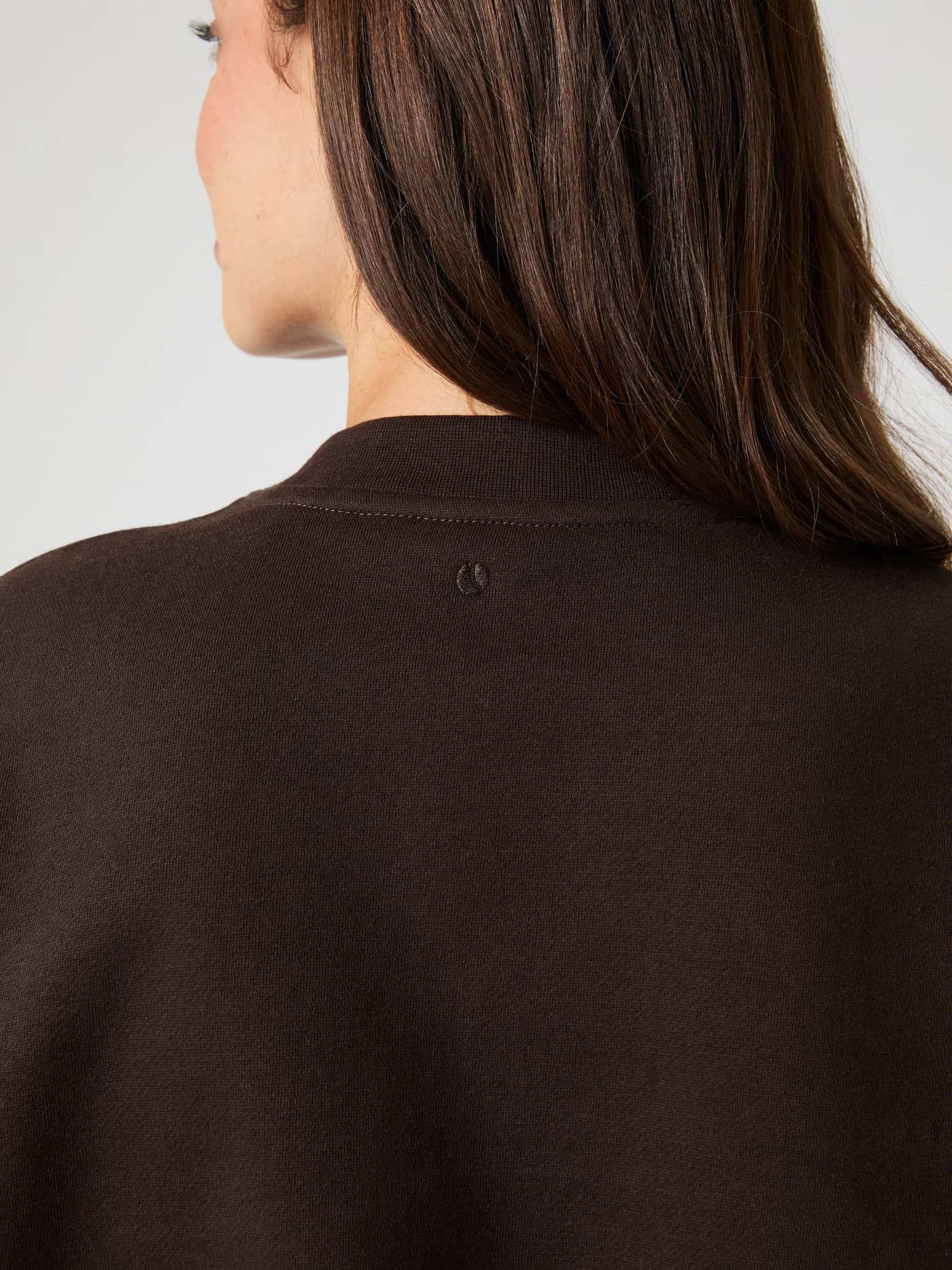 Close-up of the back of the neck of a model wearing the brown Studio Oversized Sweatshirt, showing the small logo. - Image 4
