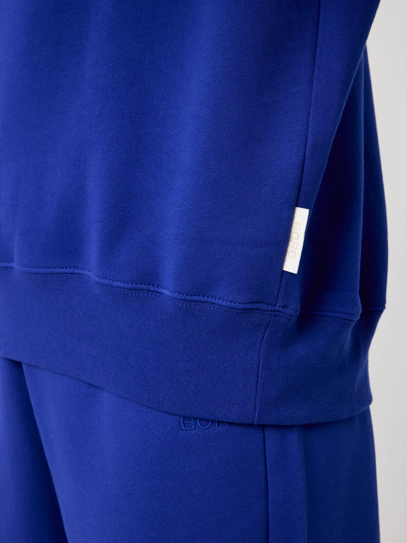 Detailed close-up of the Studio Oversized Sweatshirt in Clematis Blue (Blue) with a small white BORG label at the hem. - Image 5