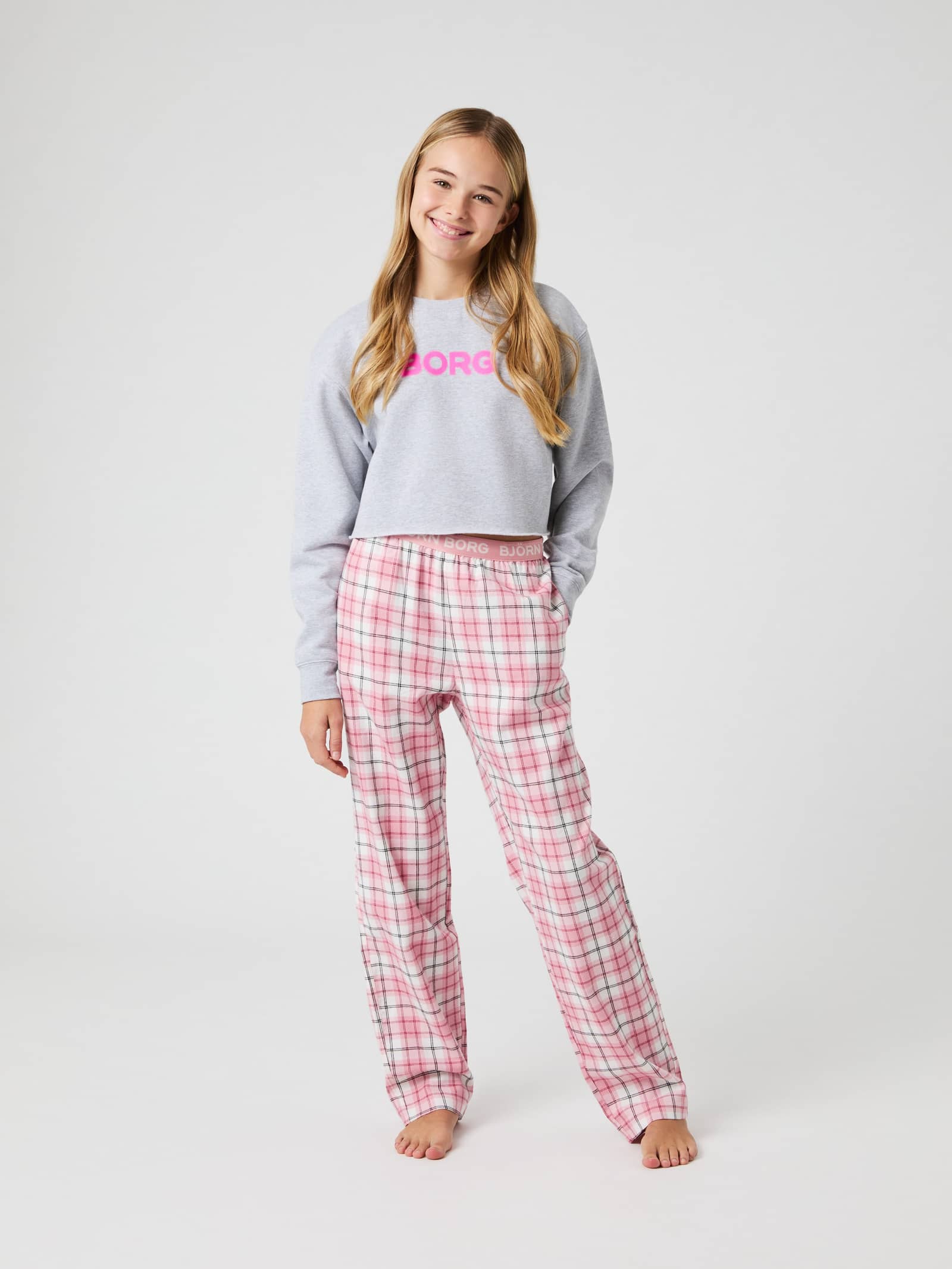 Girl smiling while wearing Björn Borg Core Flannel Pyjama Pants in a pink tartan print, styled with a grey sweatshirt. - Image 1