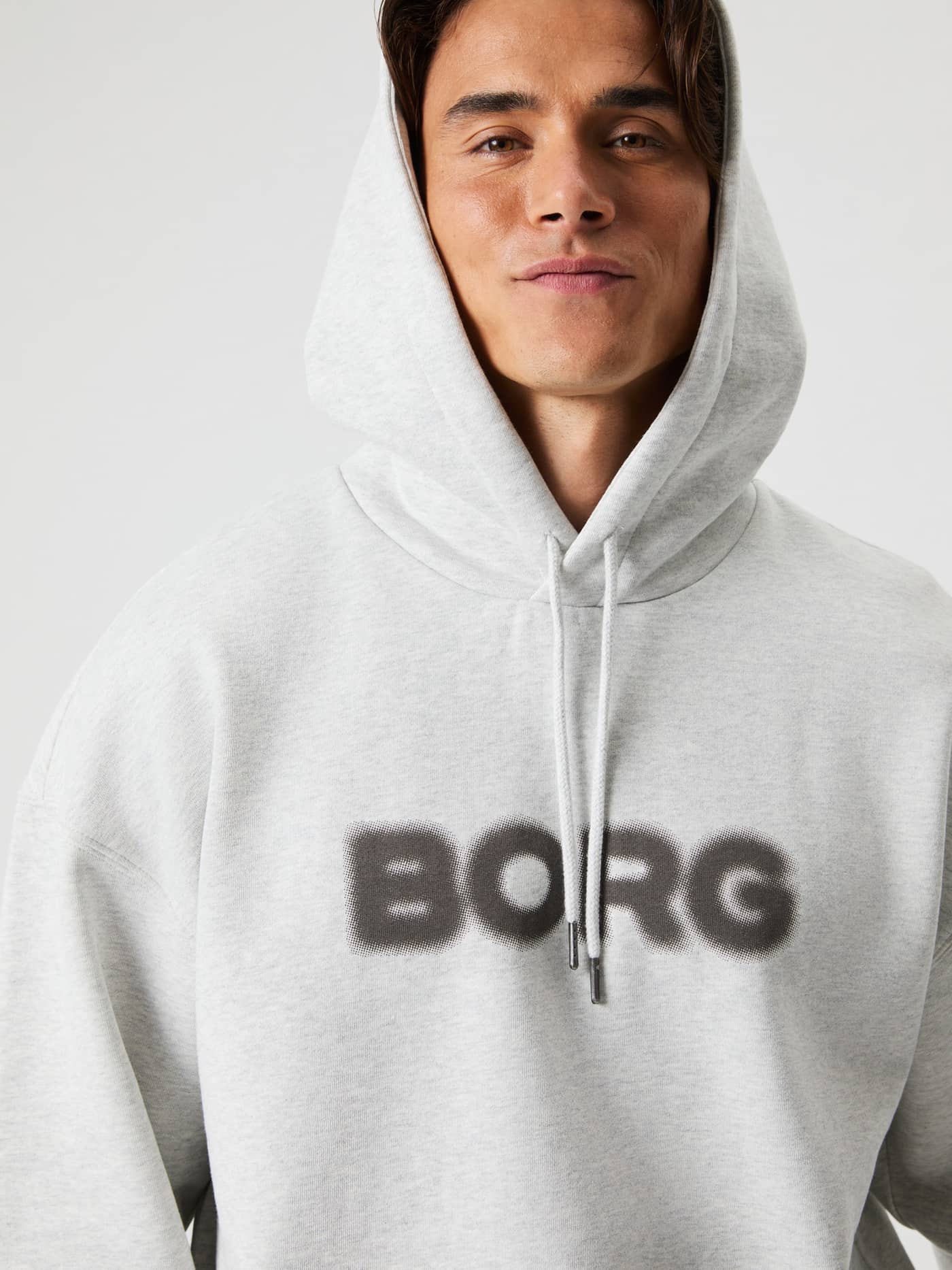 Primer plano de un hombre con la Borg Oversized Hoodie en Ultra Light Grey Melange (Gris) y logo frontal oscuro. - Image 1