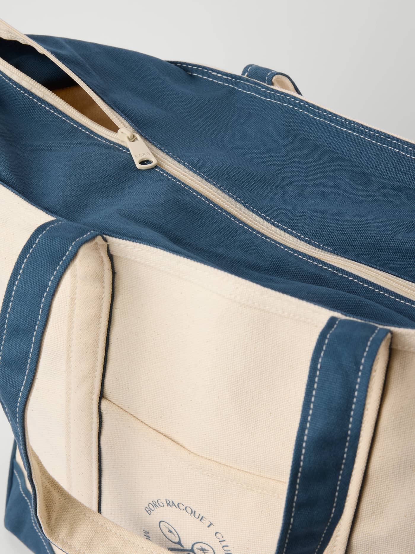 Close-up of the cotton Ace Canvas Tote 30L in Egret (White) featuring a zipper and blue Racquet Club logo for tennis. - Image 3