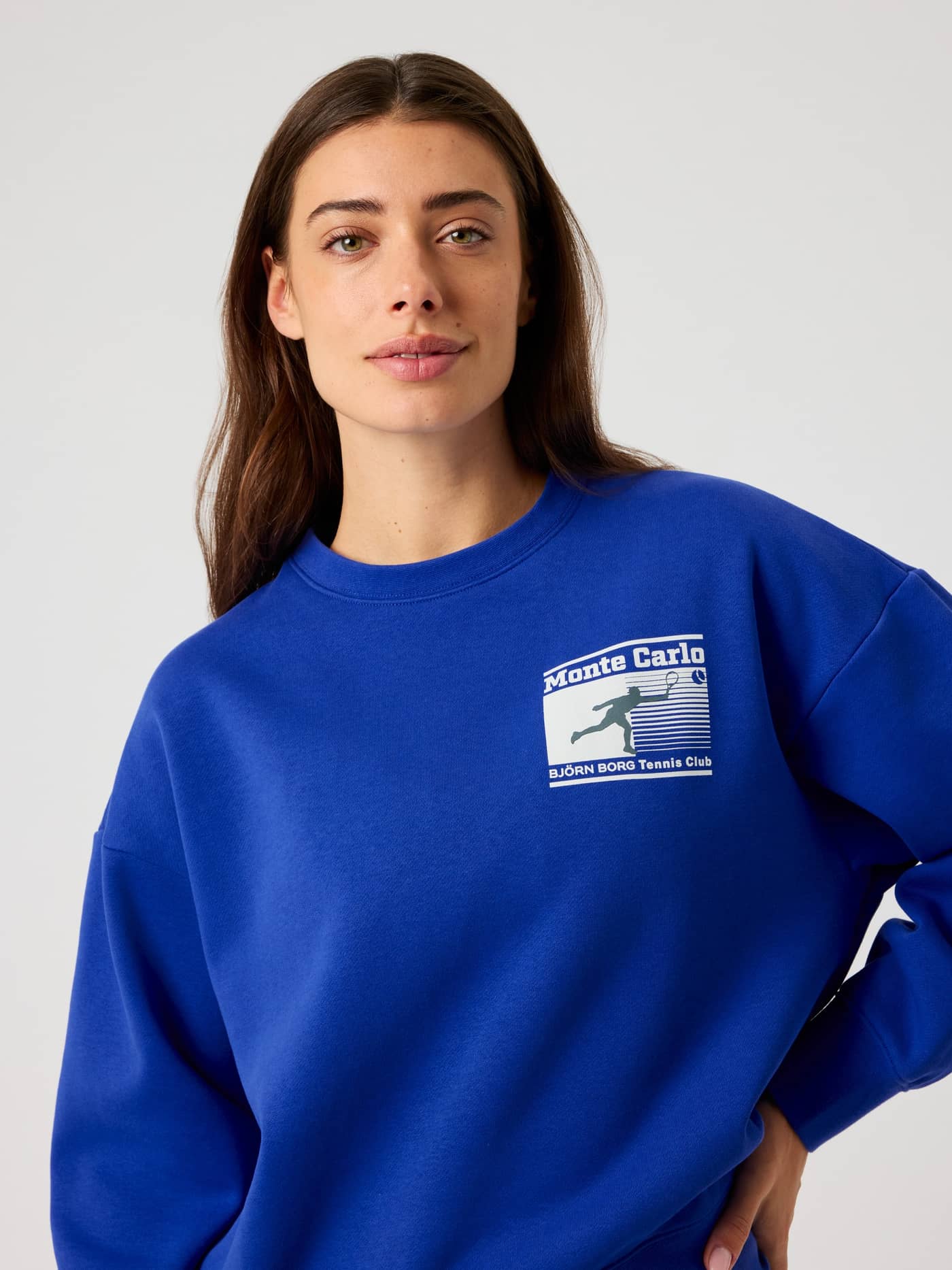 Vorderansicht einer Frau im weichen, oversized Borg Boyfriend Sweatshirt in Clematis Blue (Blau) mit weißem Tennis-Logo. - Image 6