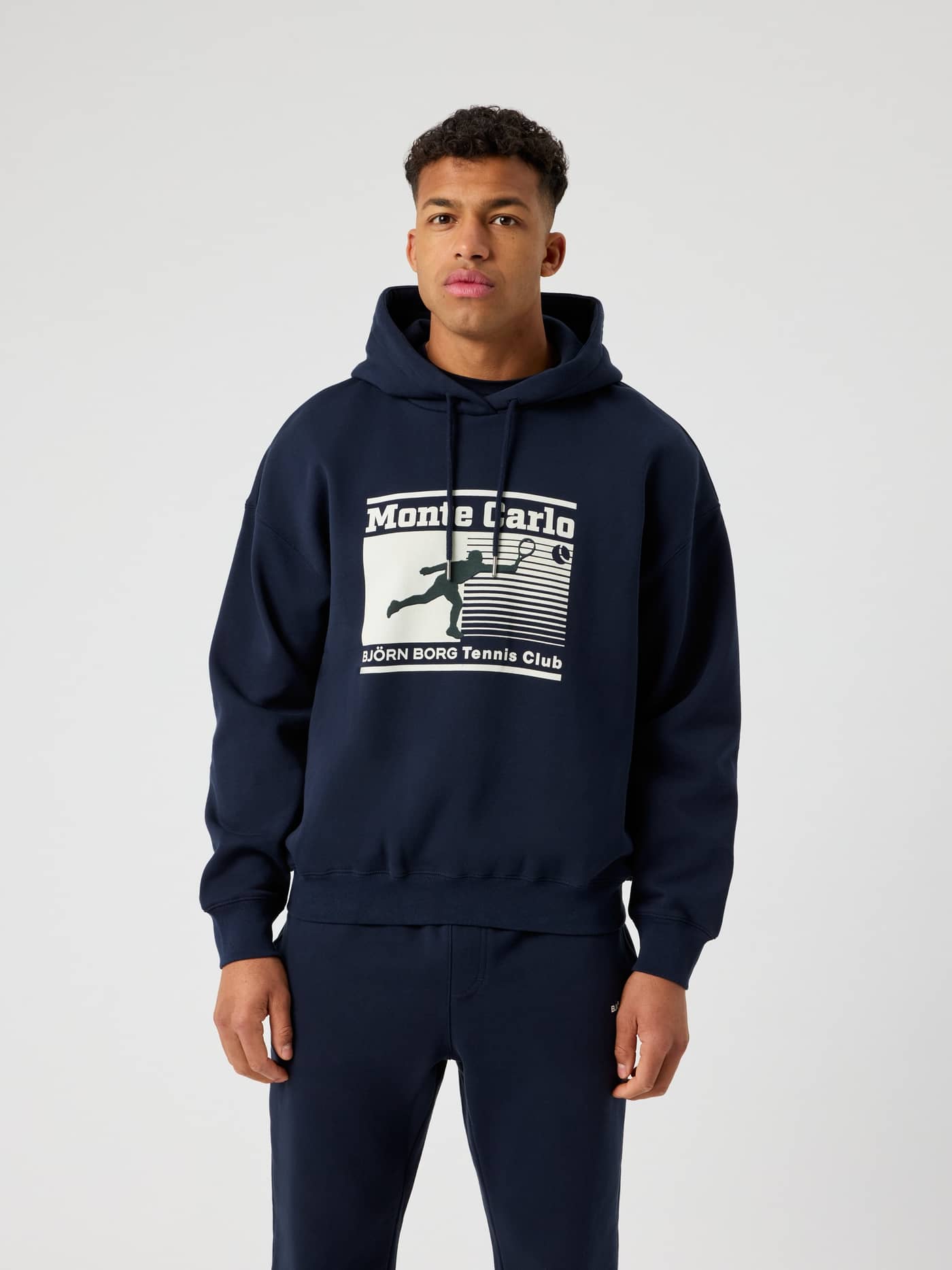 Front view of a man in the Night sky (Navy) Borg Oversized Hoodie with a Monte Carlo tennis graphic for style and function. - Image 2