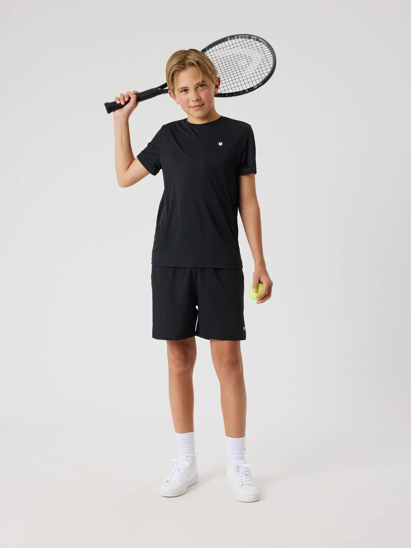 Vooraanzicht van een kind in het Black Beauty (Zwart) Ace Light T-Shirt met wit logo en een tennisracket voor kids. - Image 4