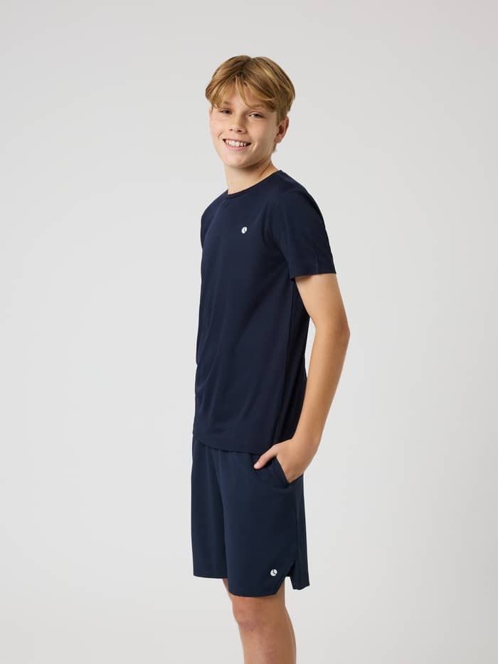Side view of a boy in the Ace Light T-Shirt in Night sky (Navy), a breathable kids' tennis top featuring a white logo.