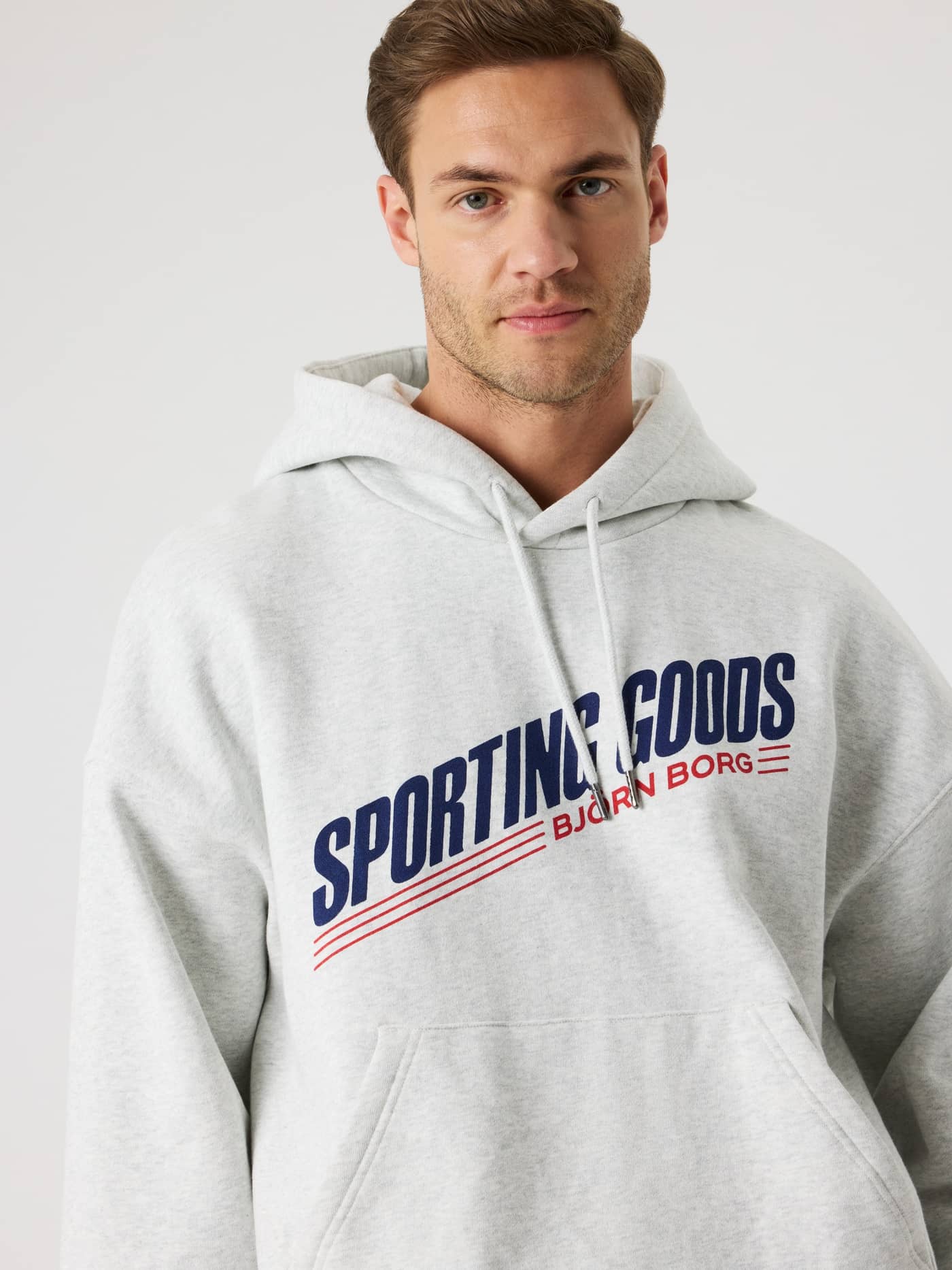 Close-up of a man in the Borg Oversized Hoodie in Ultra Light Grey Melange (Grey) with a navy and red tennis logo. - Image 8