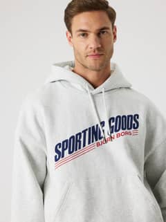 Close-up of a man in the Borg Oversized Hoodie in Ultra Light Grey Melange (Grey) with a navy and red tennis logo. - Image 8