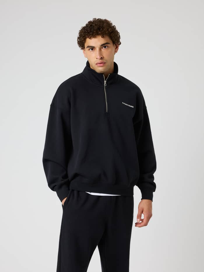 Vorderansicht eines Models im Borg Oversized Half-Zip in Black Beauty (Schwarz) mit weißem Logo zum Training.