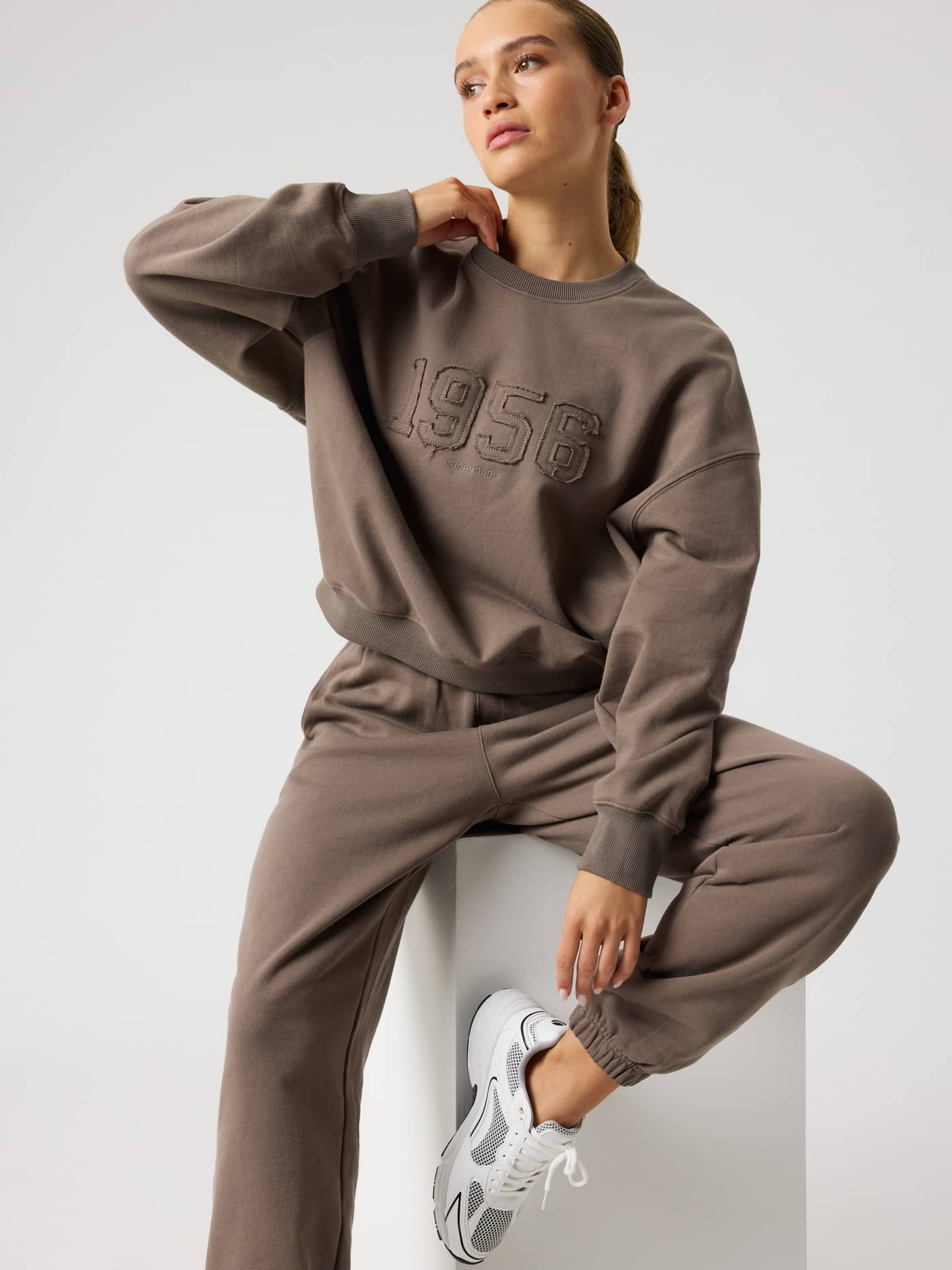 Model sitting in the Studio Oversized Sweatshirt in Falcon (Brown), showcasing a relaxed fit and 1956 chest logo. - Image 6