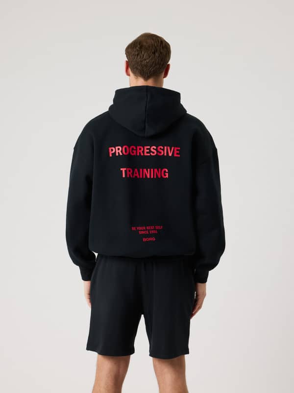 Vue de dos d'un homme portant le Borg Oversized Hoodie en Black Beauty (Noir) avec graphismes d'entraînement rouges.