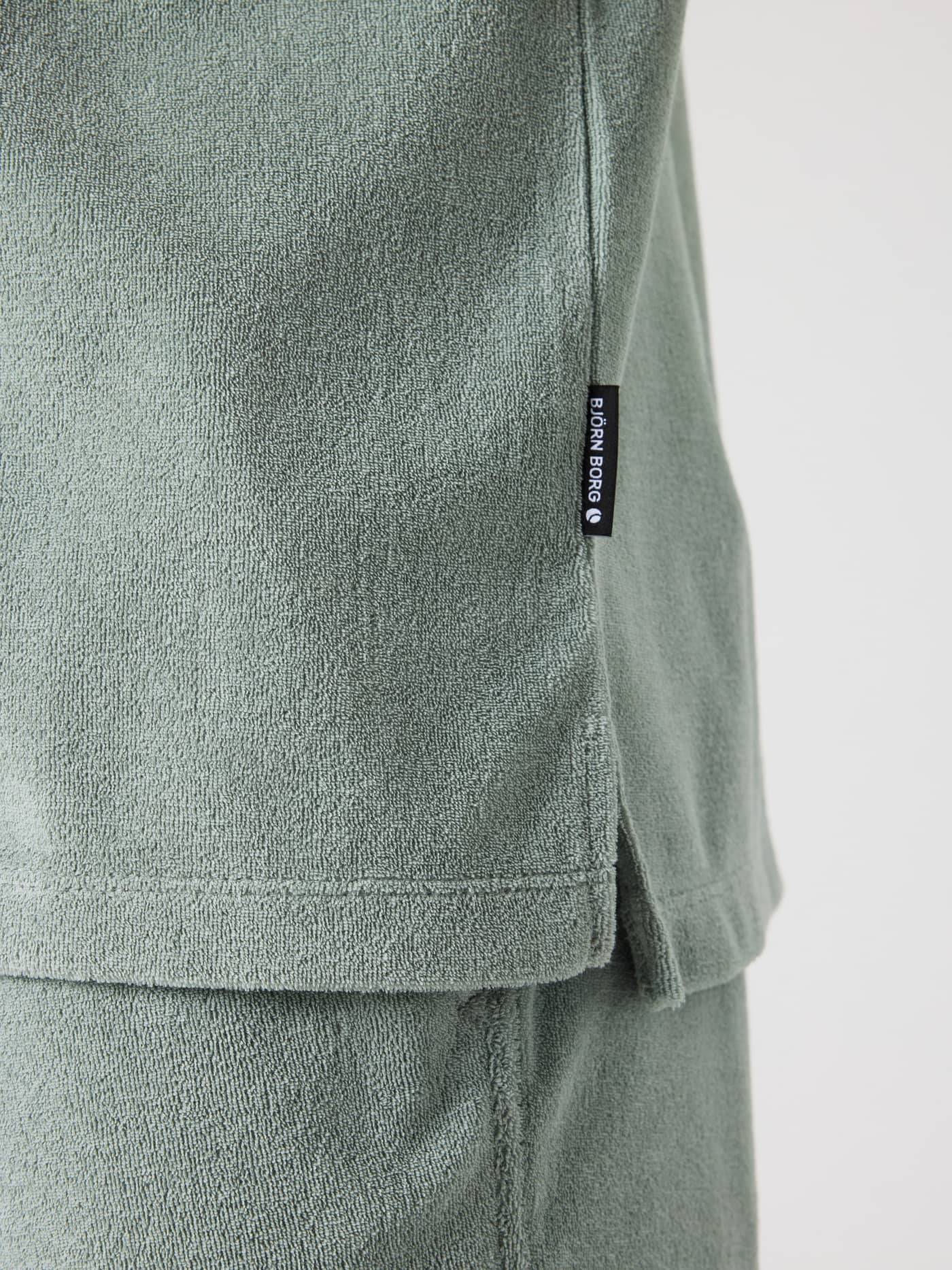 Side slit detail of the Borg Toweling Pool Shirt in Slate Gray (Green), featuring terry toweling and a black logo label. - Image 5