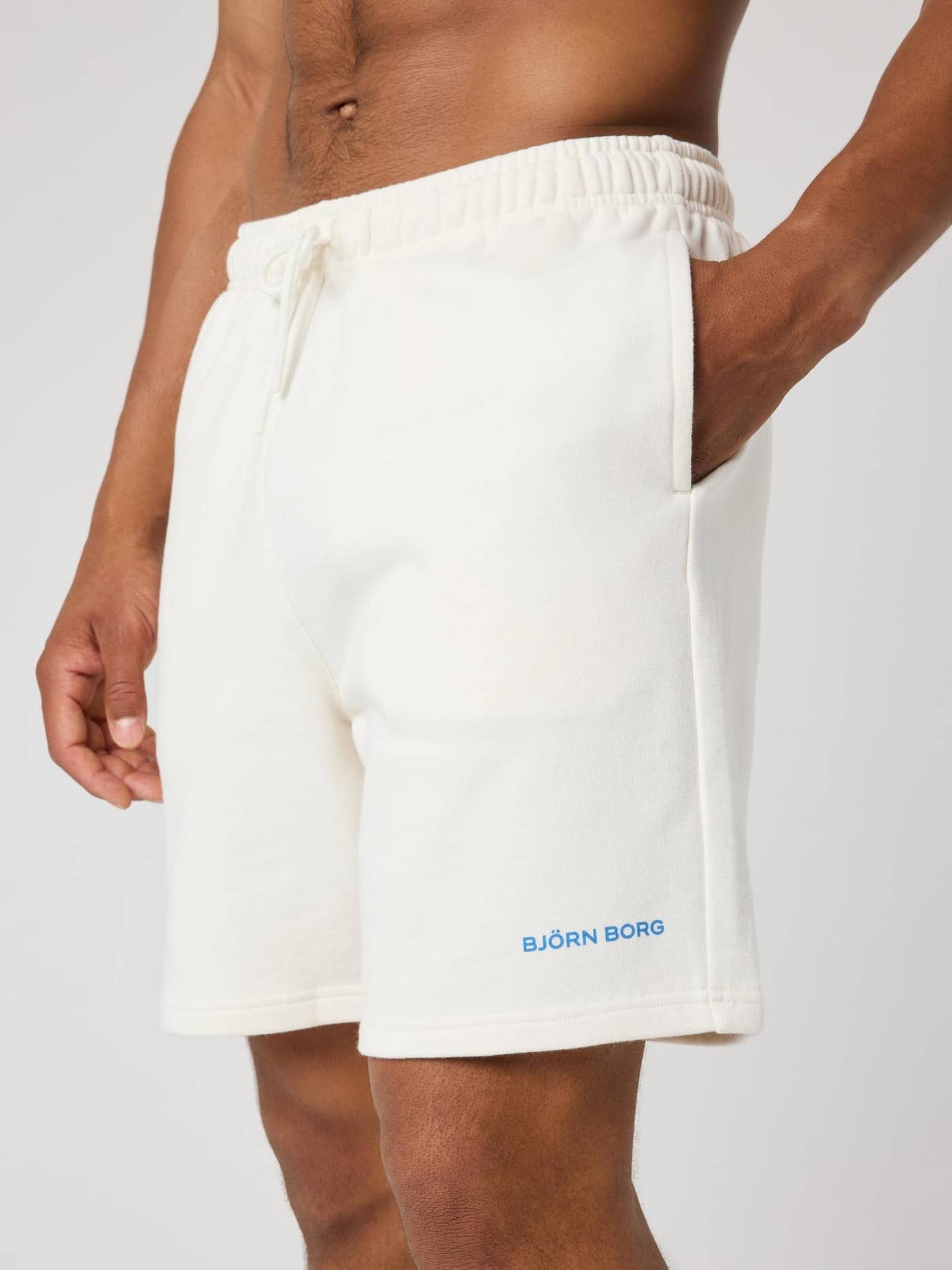 Vista lateral de un hombre con los Borg Classic Sweatshorts en Tofu (Blanco) de french terry con logo azul. - Image 4