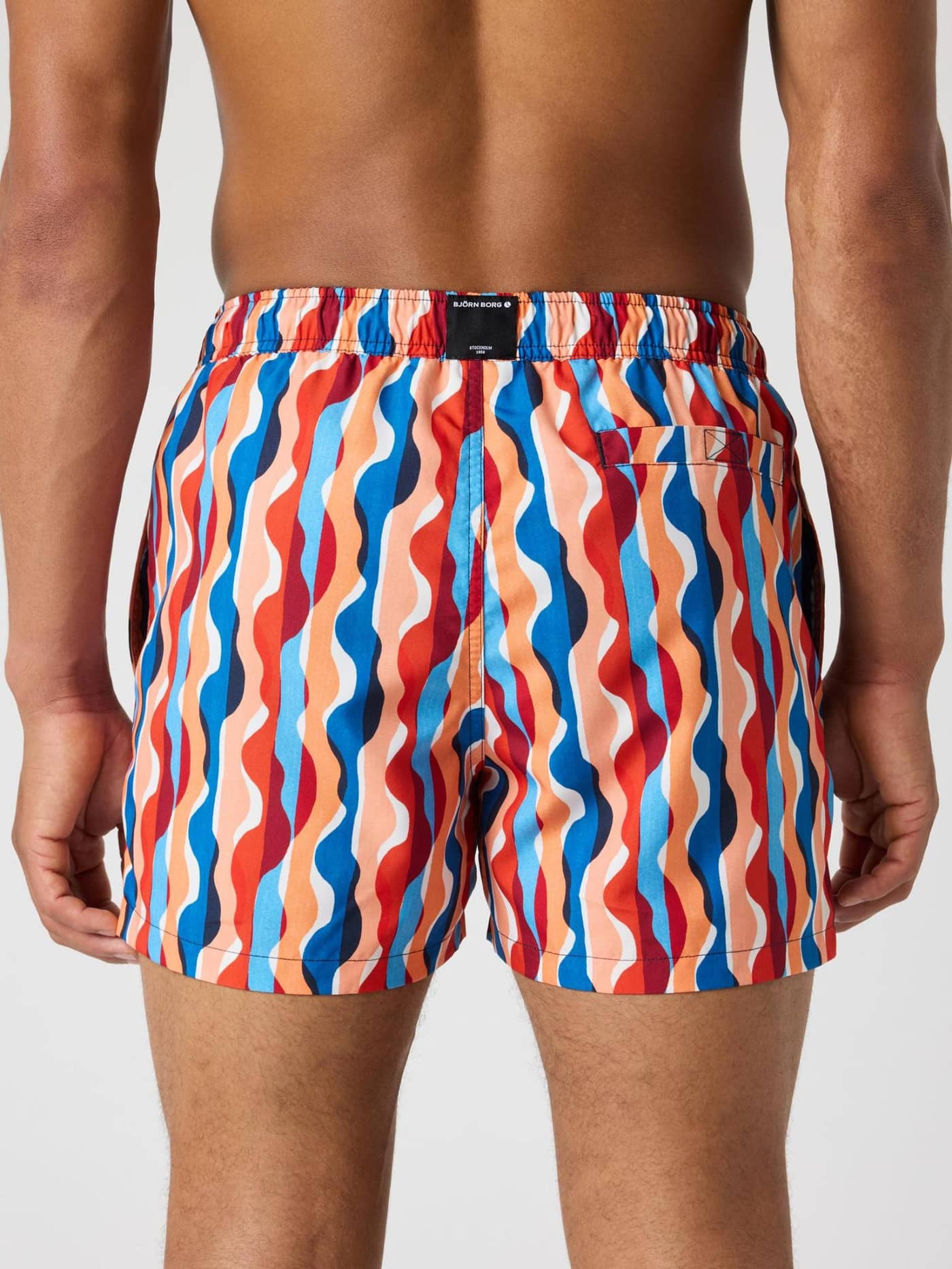 Vista posterior de los Borg Print Swim Shorts en azul, rojo y naranja con estampado digital ondulado y etiqueta negra. - Image 2