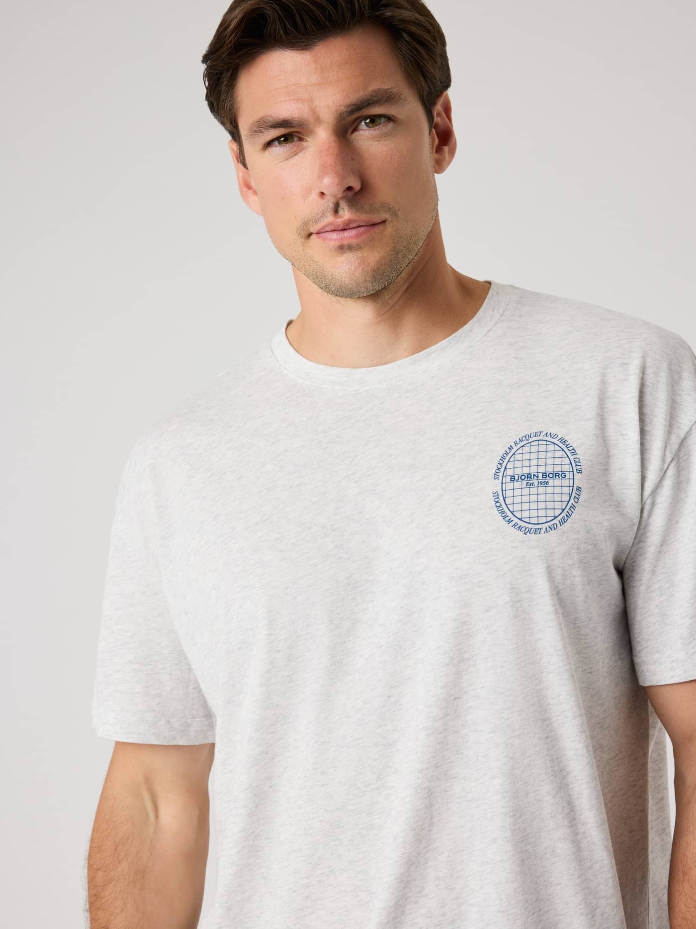 Front view of a model wearing the Ace Oversized T-Shirt in Ultra Light Grey Melange (Grey) with a blue tennis graphic. - Image 5