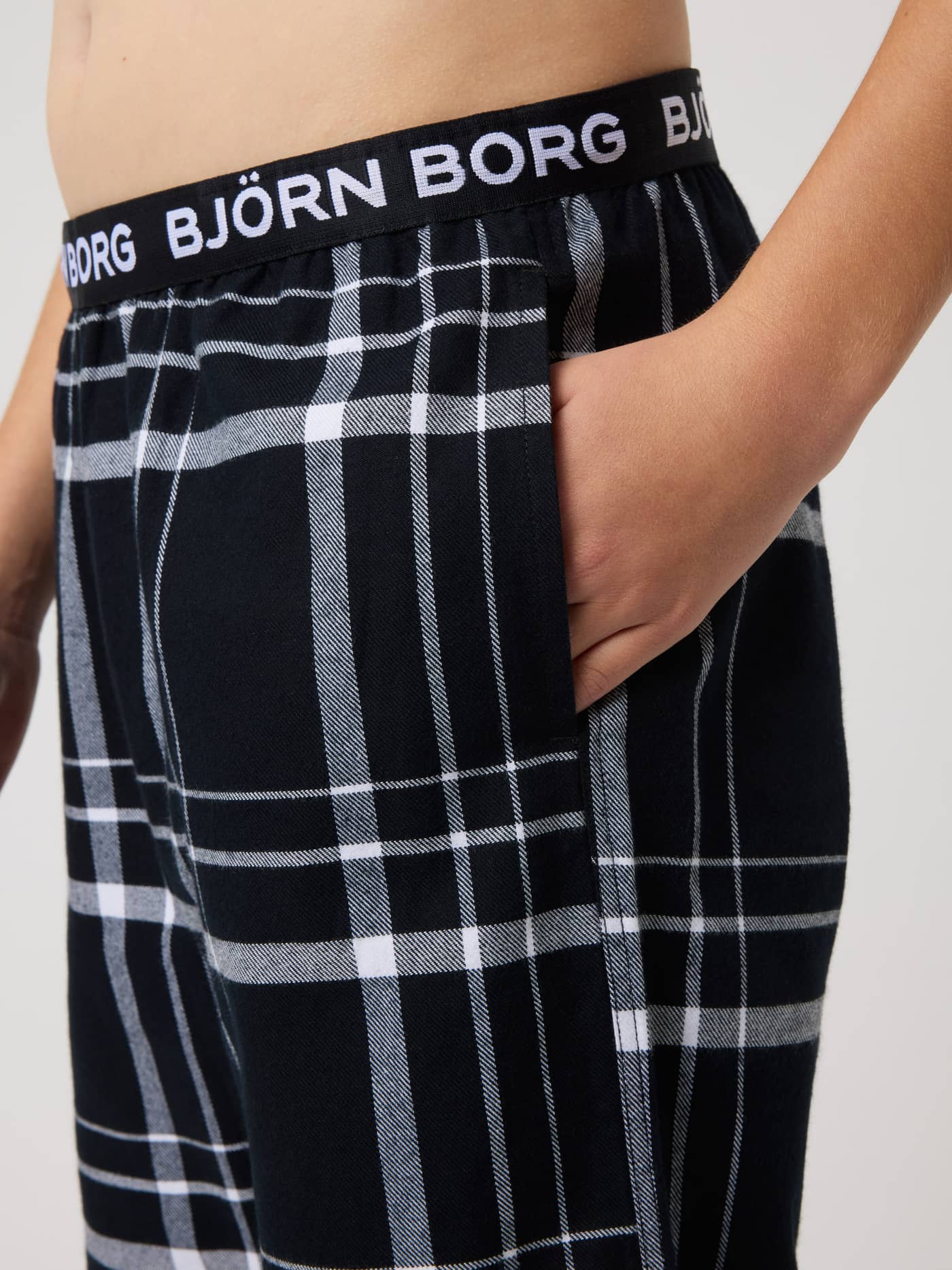 Close-up of kids Core Flannel Pyjama Pants in black, grey, and white checks, featuring a white logo on the waistband. - Image 4