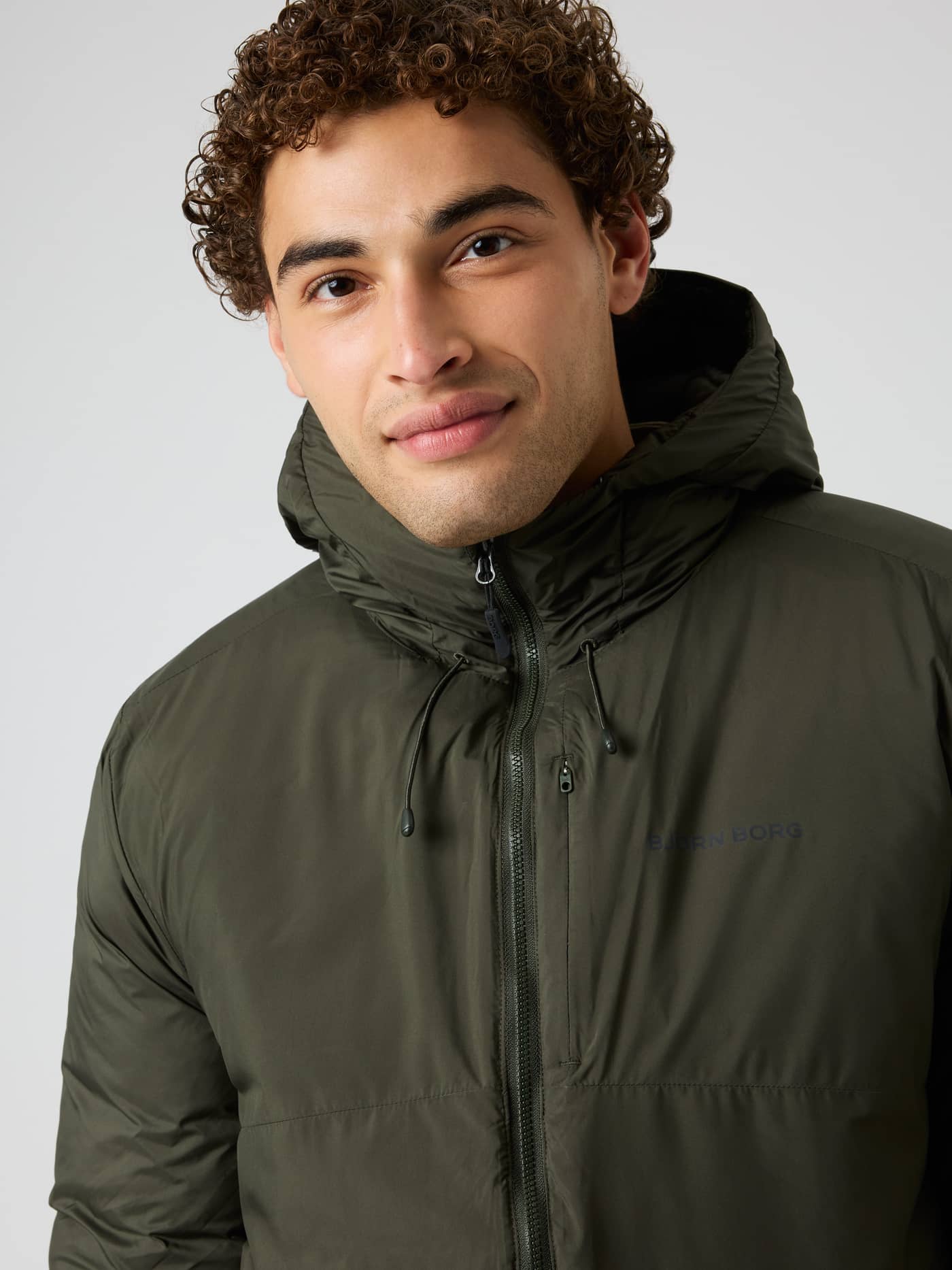 Nærbilde av vattert Borg Liner Hood Jacket i Rosin (Grønn) for trening og tennis med en mørk brystlogo. - Image 4